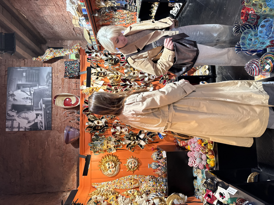 The image captures an interior view of a retail shop in Venice, Italy, specializing in Venetian masks and glass art. The shop features walls partially covered in exposed reddish-brown brick and vibrant orange panels. A large black and white photograph depicting two men, likely artisans, working in a workshop is mounted on the brick wall. The orange wall panels are densely covered with various Venetian carnival masks, including elaborate full-face masks, sun-themed designs, and jester masks.
Shelves and display cases throughout the shop present numerous glass art objects, such as colorful vases, swirl-patterned spheres, and a tall, abstract sculpture composed of multi-colored glass fragments. A large purple bowl and a red and wood oval dish are visible on an upper shelf.
Two adult females are present, both appearing to browse the merchandise. The woman on the left, with long brown hair, wears a long, light beige trench coat and is positioned facing the mask display on the orange wall. The woman on the right, with short light blonde hair, wears a light brown jacket, a lighter-colored top, and light blue jeans, carrying a dark brown leather messenger bag across her body. Her head is tilted downwards, suggesting she is examining items on a lower display or shelf. The floor is dark and reflective.