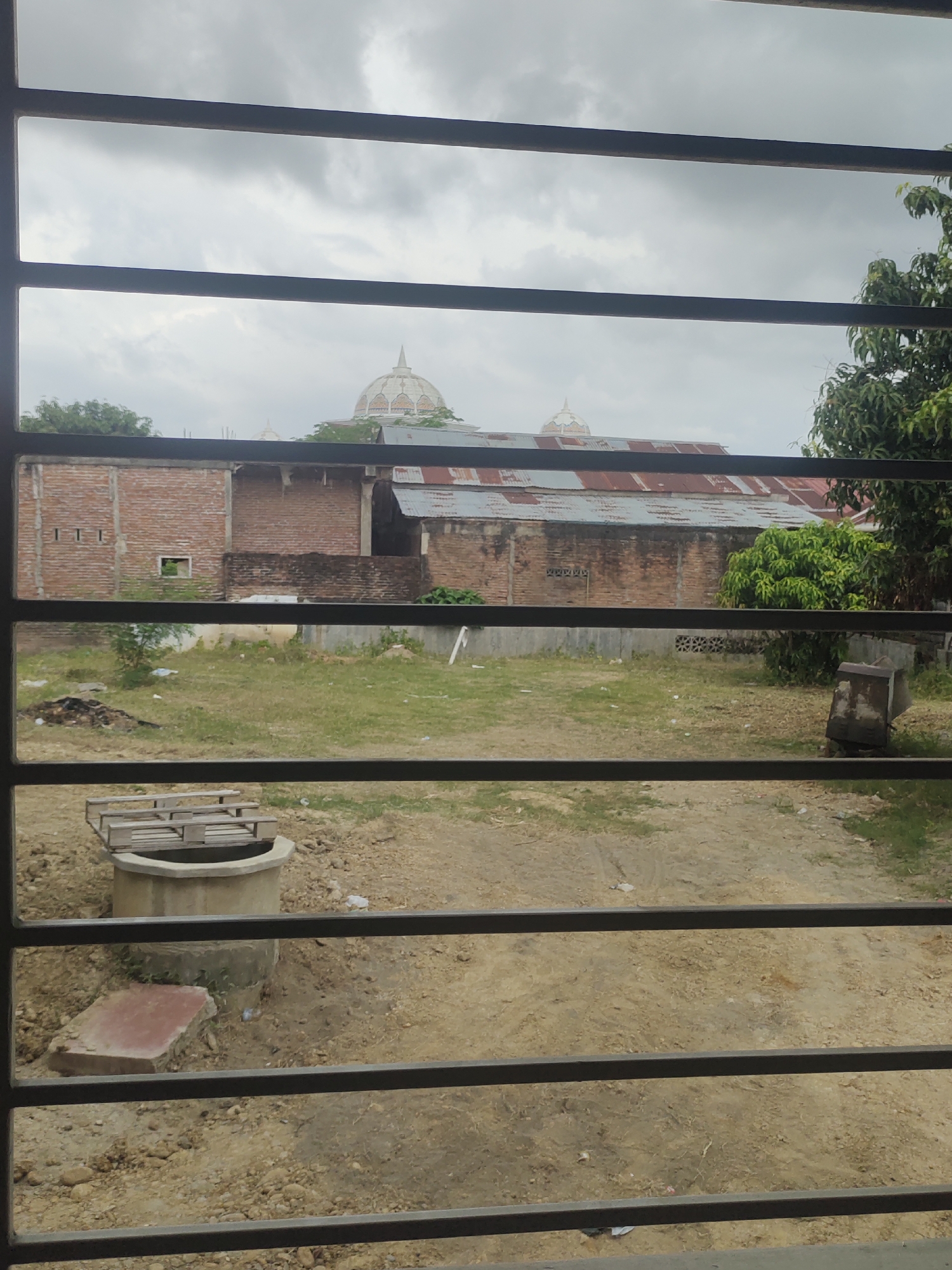 The image captures an outdoor scene viewed through a window or security bars, evident from the dark horizontal metallic stripes in the foreground. The setting appears to be a semi-rural or undeveloped area, with the provided context placing it in Meunasahaluedrienlhoksukon Barat, Indonesia.
In the immediate foreground, a concrete well, covered with a wooden pallet, is visible, alongside a reddish rectangular concrete slab. The ground beyond the bars consists mainly of bare earth with scattered patches of dry grass and some debris.
Further into the midground, an unkempt plot of land stretches out, featuring areas of sparse grass and dirt. To the left, a brick building with small rectangular ventilation openings is partially visible. Adjacent to it, another brick structure dominates the midground with a rusty, corrugated metal roof. A smaller building with a similar roof is seen towards the right, partly obscured by lush green trees and bushes that dot the landscape.
In the background, under a heavily overcast and cloudy sky, two prominent domed structures with small spires, likely mosques, rise above the rooftops. These structures feature light-colored, ornate details.
The scene conveys a sense of stillness; no people or specific activities are observed. The cloudy sky suggests an overcast day, possibly indicating a lack of direct sunlight or impending rain. There is no visible text in the image.