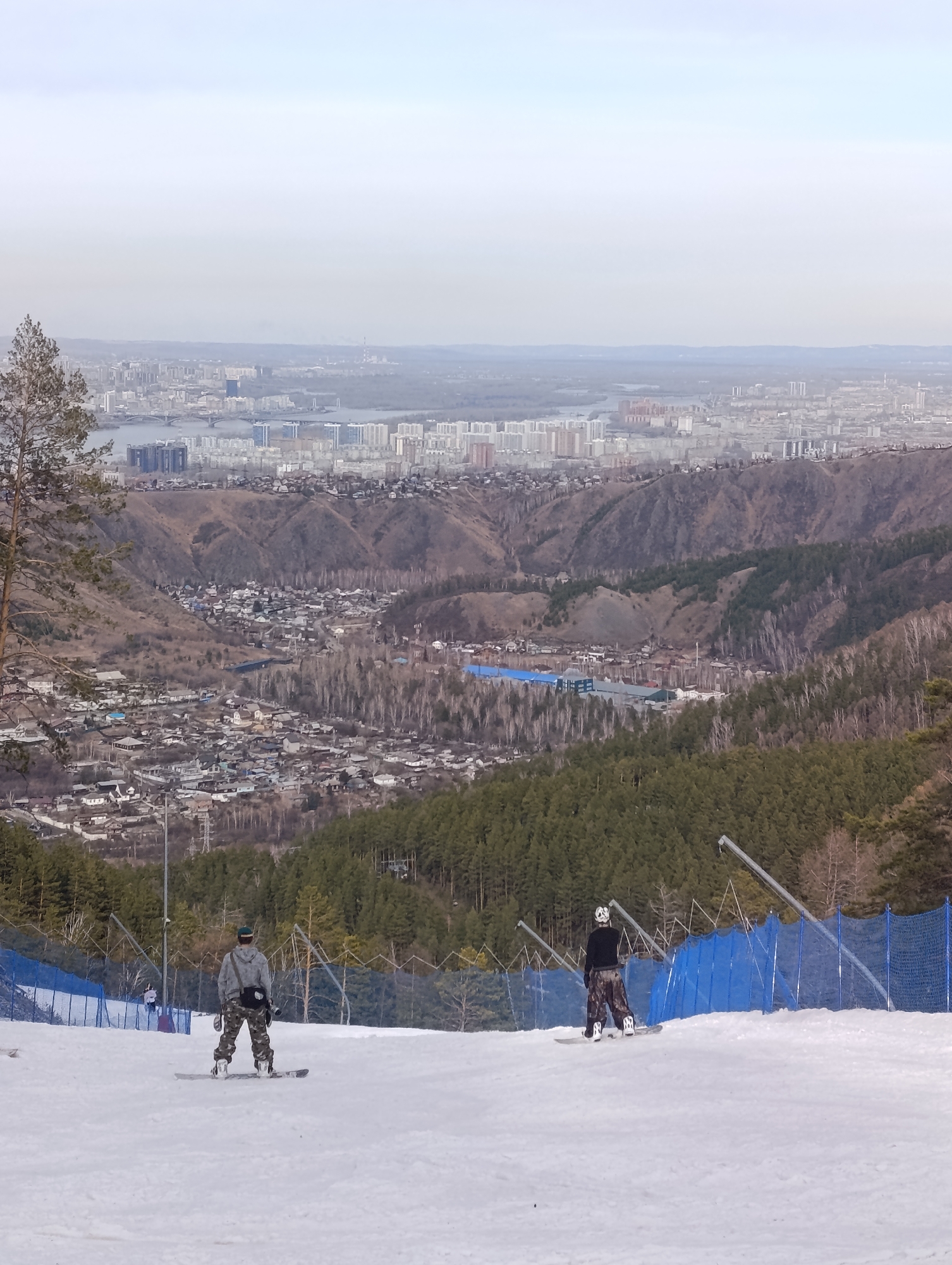 The image depicts two snowboarders standing on a snow-covered slope, facing away from the viewer. A blue safety fence runs along the side of the slope. Behind the slope is a densely wooded area. Beyond the trees, a cityscape extends into the distance, partially obscured by haze or smog. This area is Krasnoyarsk, Russia. The terrain consists of hills and valleys. The sky is overcast and light in color.