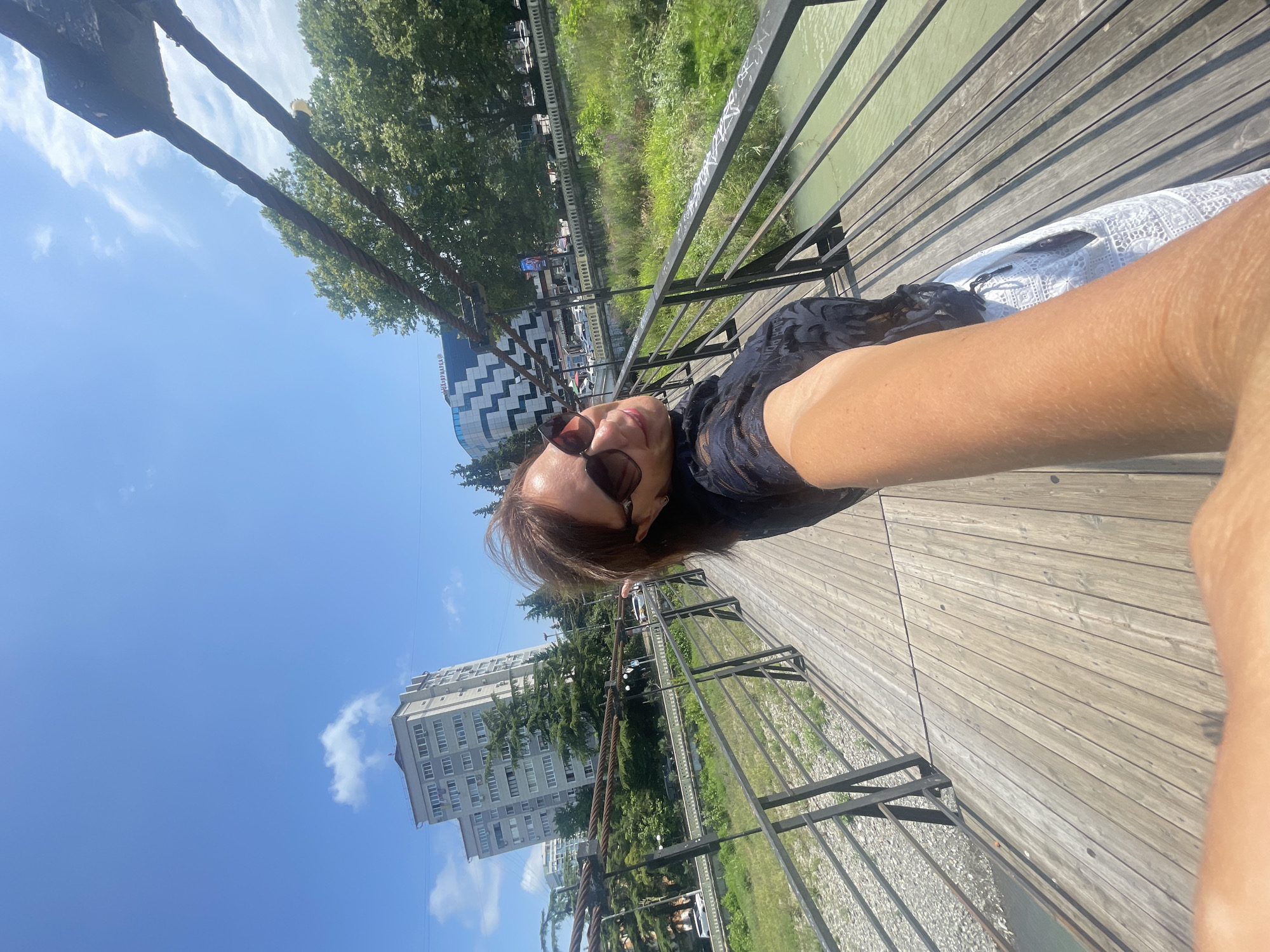 A woman is standing on a pedestrian bridge, holding her phone to take a selfie. The bridge is made of wood and has metal railings. She is wearing sunglasses and a dark, patterned top with white, lacy pants. In the background, there is a river, green vegetation, and buildings. To the left, a tall, light-colored building with many windows stands against a clear blue sky with a few white clouds. Trees line the riverbank. The architecture suggests a location in Sochi, Russia. The overall impression is a sunny day with a relaxed atmosphere.