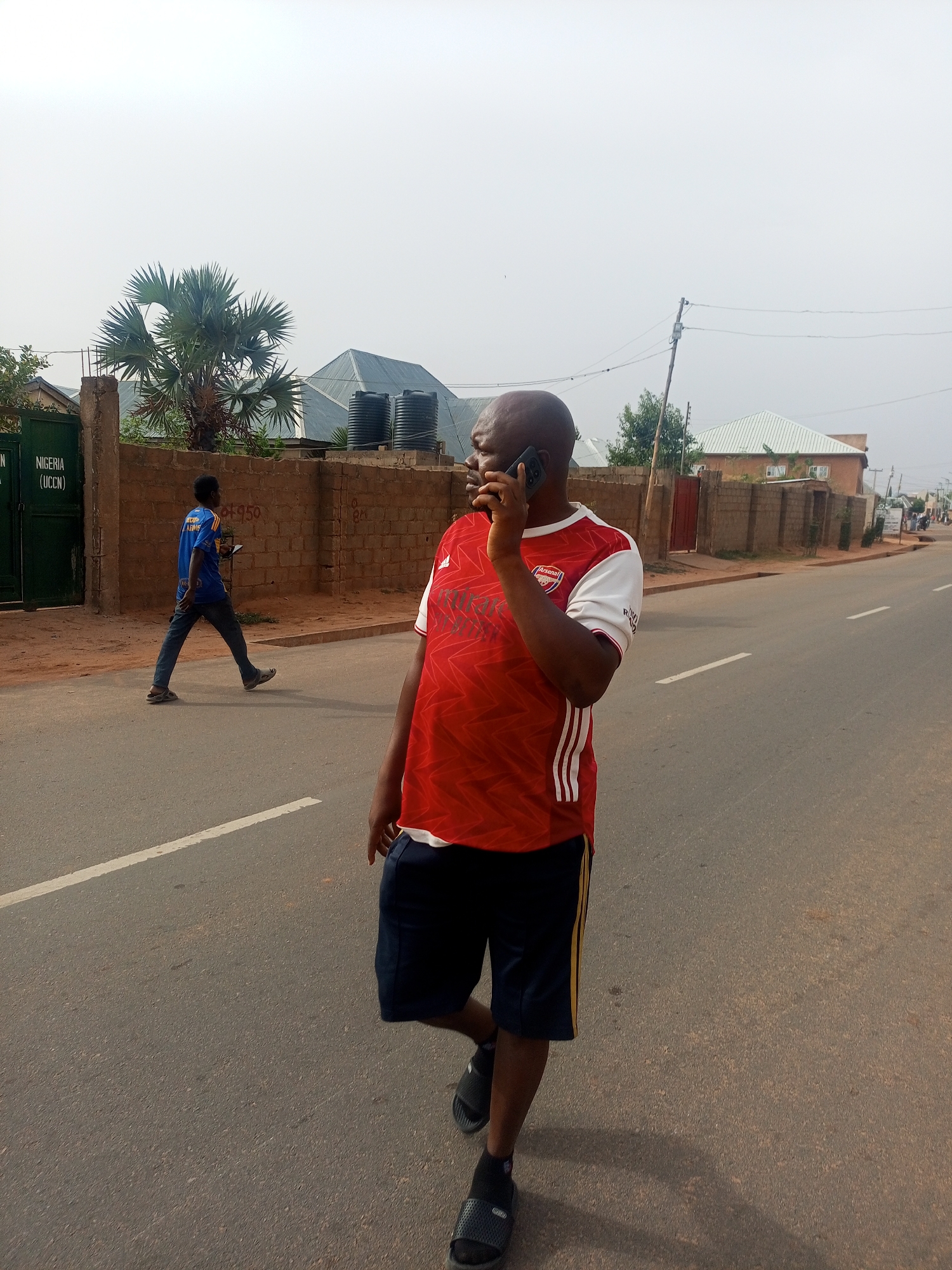 A man wearing an Arsenal soccer jersey and dark shorts is walking on a paved road while talking on a smartphone. The street is lined with brick walls and buildings, some with water tanks on the roofs. Another man is walking on the same side of the road in the distance. The environment appears to be a city in Nigeria, specifically Tempure, Nigeria, based on the writing on the gate on the left. A palm tree is visible behind the brick wall.