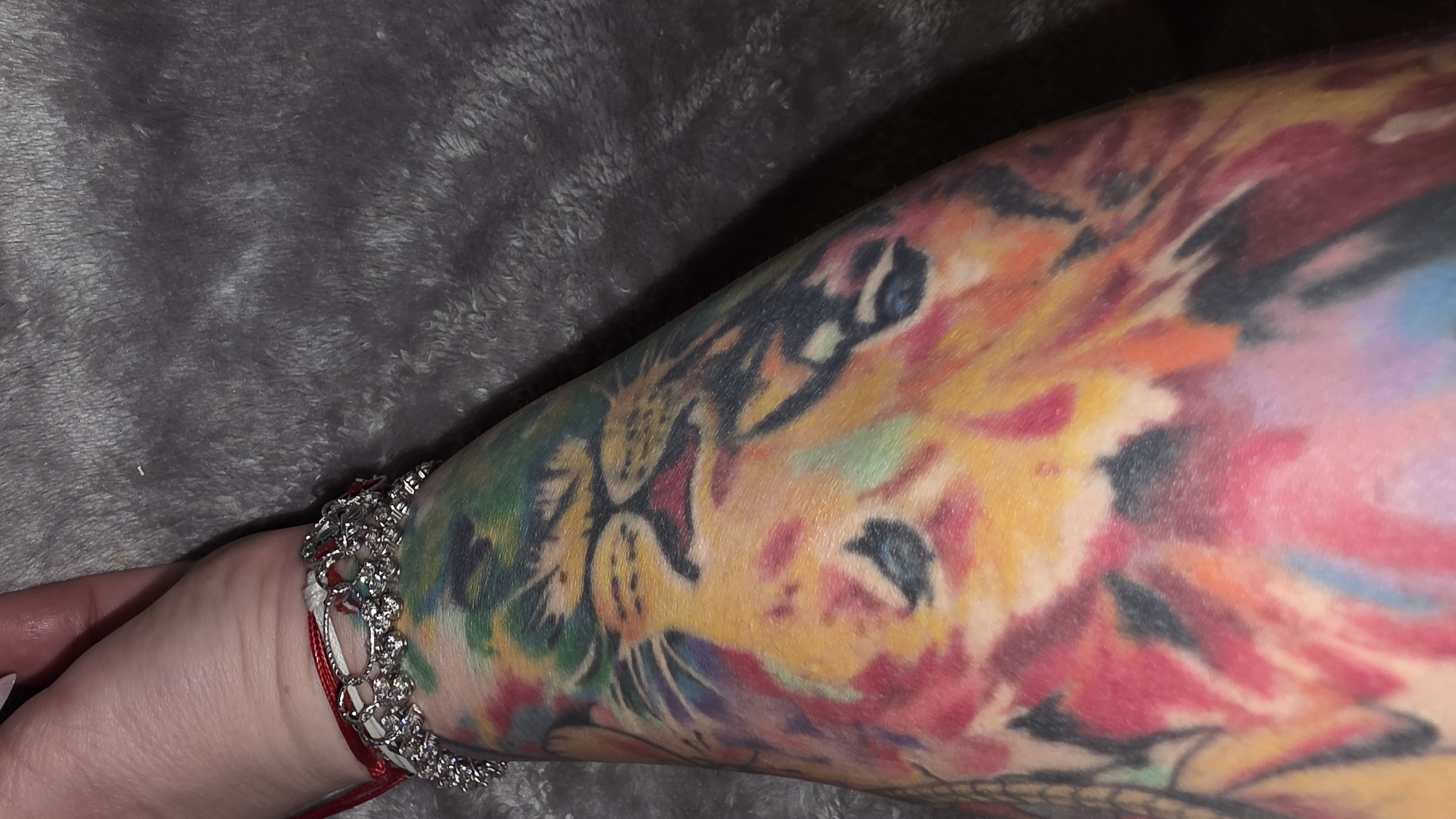 A close-up of a person's forearm displaying a colorful tattoo of a lion's head. The tattoo incorporates a variety of colors including yellow, red, green, and blue. A bracelet with silver-colored, rhinestone-like elements and a red string is around the wrist. The forearm is resting against a soft, grey, textured surface.