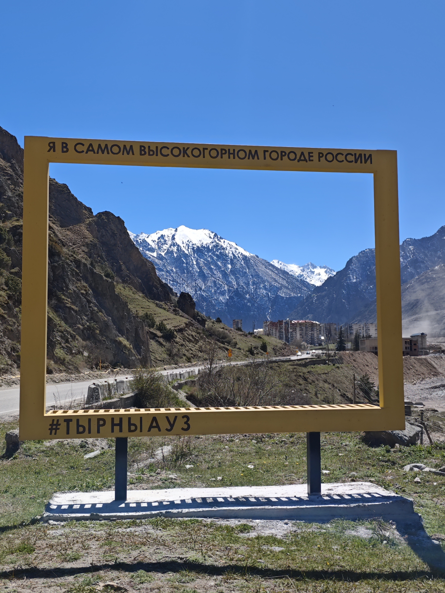 A medium shot depicts a yellow metal frame, partially enclosing a view of mountains and the town of Tyrnyauz, Russia. The frame, with text in Russian, is grounded on a concrete base with two supporting columns. The landscape behind the frame includes a winding road, mountain slopes, and residential buildings clustered in the valley. Snow-capped peaks rise in the distance under a clear blue sky. The foreground consists of a patch of low-lying grass and vegetation, and evidence of construction or excavation is visible near the town. The overall scene conveys a sense of place, framing the natural and built environment of Tyrnyauz.