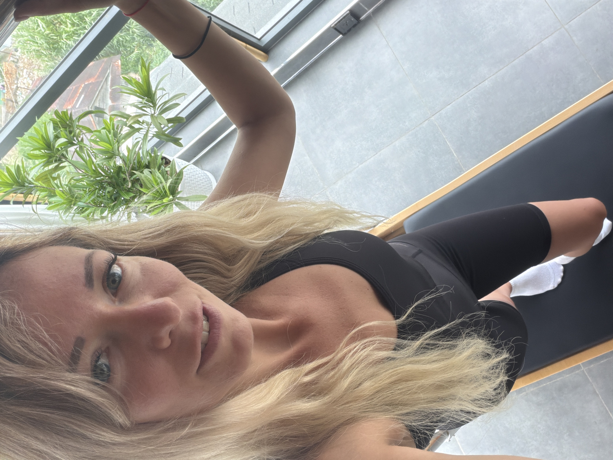 A medium shot captures a blonde woman indoors, possibly in Da Lat, Vietnam, looking directly at the camera with a slight smile. She is positioned on a Pilates reformer, a piece of exercise equipment. The woman wears a black sports bra and black cycling shorts, with white socks on her feet. Her long, wavy blonde hair frames her face. In the background, a large window reveals a lush green plant in a white pot on a windowsill, and beyond that, a glimpse of a building with a reddish-brown roof and dense foliage, suggesting an outdoor scene. The floor is tiled in a light gray. The woman's right arm is extended upwards, grasping a window frame, and she is wearing thin black and red bracelets on her wrists. The lighting appears natural, coming from the window.