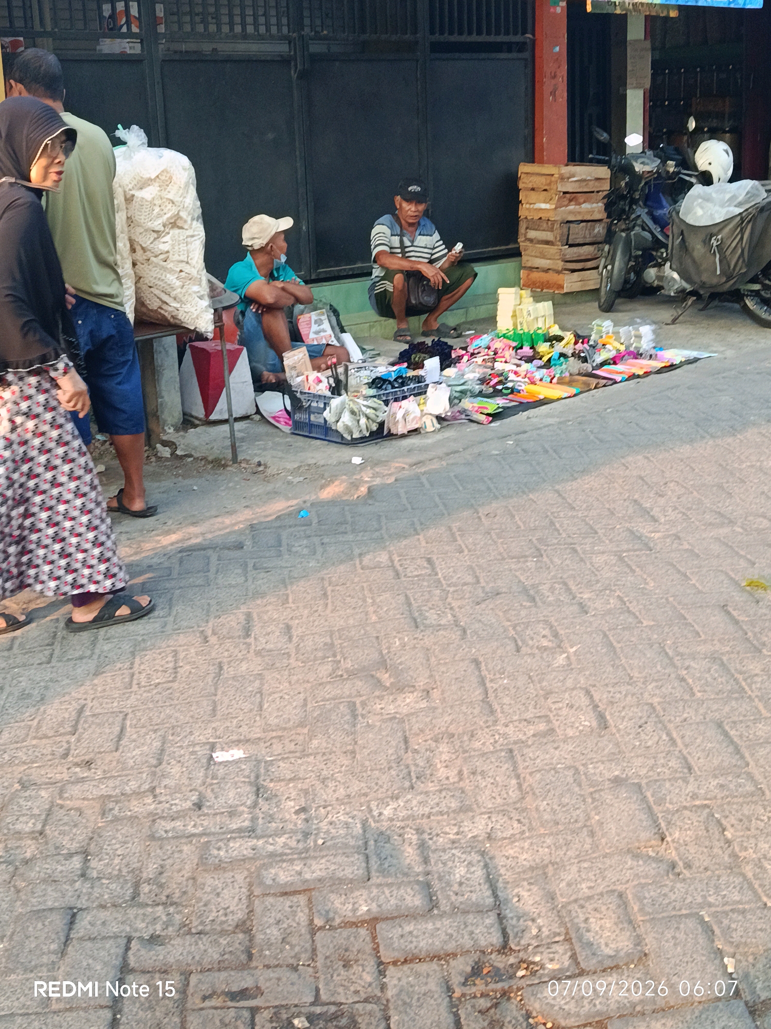 The image captures a street scene in Keboansikep, Indonesia, likely in the morning given the timestamp. A woman wearing a black hijab and a patterned skirt walks from the left side of the frame. Next to her is a person in a green shirt and blue shorts, and a large bag of white puffed snacks. Two men are seated on the ground in the middle of the frame, engaged in what appears to be selling or displaying goods. One man, wearing a light blue shirt and a beige cap, is looking towards the left. The other man, in a striped shirt and dark shorts, is holding an item and looking down. In front of them is a colorful assortment of items laid out on a mat, including toys, toiletries, and other small merchandise. A motorcycle is parked on the right side of the frame, along with stacks of wooden crates. The ground is paved with brick. The overall atmosphere suggests a local market or street vendor setup.
