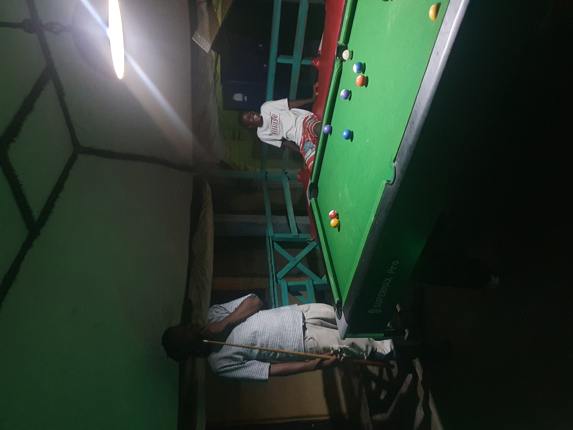The image depicts two young men playing pool in an outdoor setting, possibly a bar or entertainment area, at night. The scene is lit by a single overhead lamp, casting shadows and creating a somewhat dim atmosphere. One man is standing on the left, holding a pool cue and appears to be observing the game. He is wearing a checkered shirt and light-colored pants. The second man is seated on a bench to the right of the pool table, wearing a white t-shirt and patterned shorts, and is looking away from the camera with a smile. The pool table is green with several balls scattered across it. The background reveals a rustic structure with a sloped roof and railing. The location is identified as Lokoja, Nigeria, suggesting a vibrant local leisure spot.
