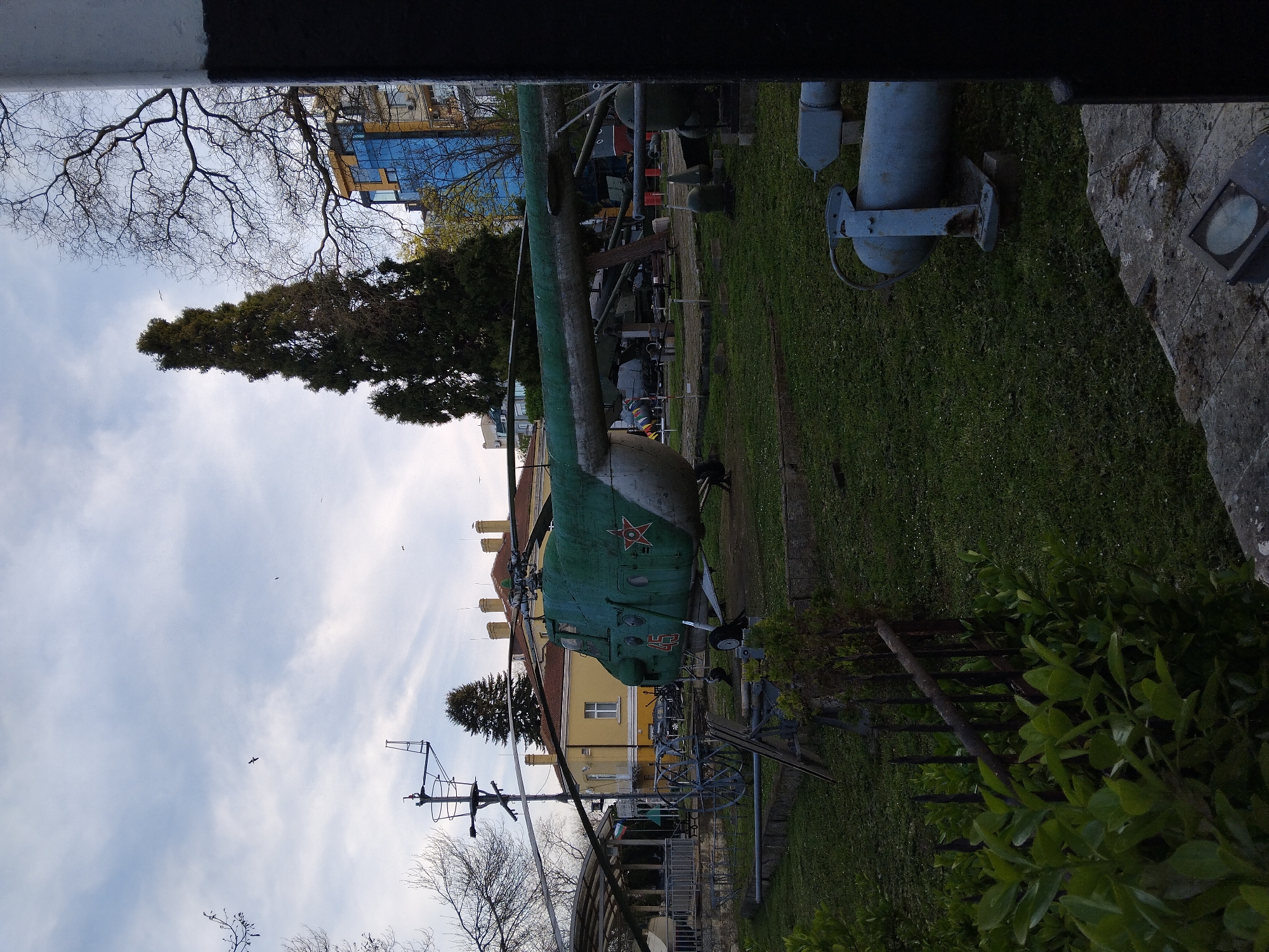 The image shows a Soviet-era Mi-24 Hind helicopter gunship on display in a grassy area at the Naval Museum in Varna, Bulgaria. The helicopter is green with a red star on its side. It is partially obscured by trees and other objects. In the background, there are buildings, including one with a blue glass facade. The foreground contains trees, bushes, and a black and white painted wall or structure partially obscuring the view. The sky is visible with some clouds and birds flying.