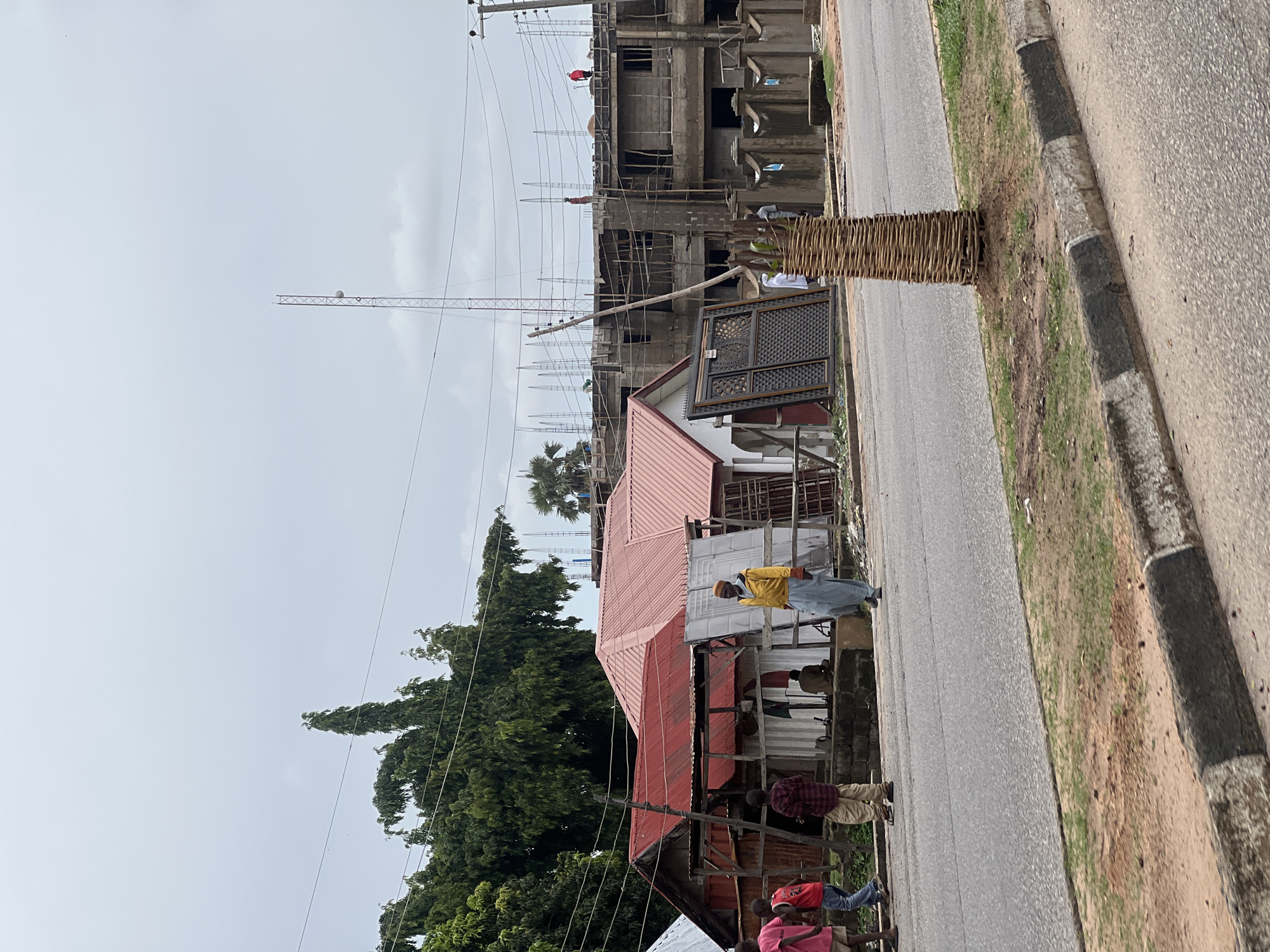 In Jalingo, Nigeria, an overcast sky looms above a street scene featuring construction and existing structures. On the left, a row of single-story buildings with red corrugated metal roofs are visible. In front of these buildings, several children and a man in a yellow jacket and blue trousers walk along the side of the road. One child wears a red shirt, another a blue shirt with "24" on it, and a third a checkered shirt and tan pants. Further to the right, a multi-story building under construction is in progress, with rebar protruding from concrete levels. Workers are visible on the upper floors, and a tall communications tower stands in the background. A concrete barrier with decorative patterns separates the construction site from the road, and a woven wooden structure encircles the base of a small tree on the roadside. Electrical wires are strung across the scene, supported by utility poles.