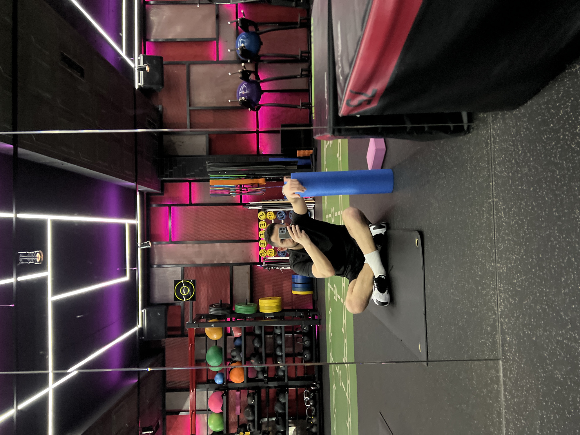 A person is reflected in a large mirror in a fitness facility, with the reflection showing the individual squatting on a grey exercise mat, holding a blue foam roller. The person is wearing a black top and shorts with white socks and black and white shoes. The mirror's reflection reveals fitness equipment, including racks with weights, medicine balls, and kettlebells. The gym has a modern design with red and grey paneled walls illuminated by pink and purple LED lighting fixtures. The gym has grey flooring with what appears to be a green turf section. The location is most likely in Sofia, Bulgaria, as that detail was provided with the prompt.