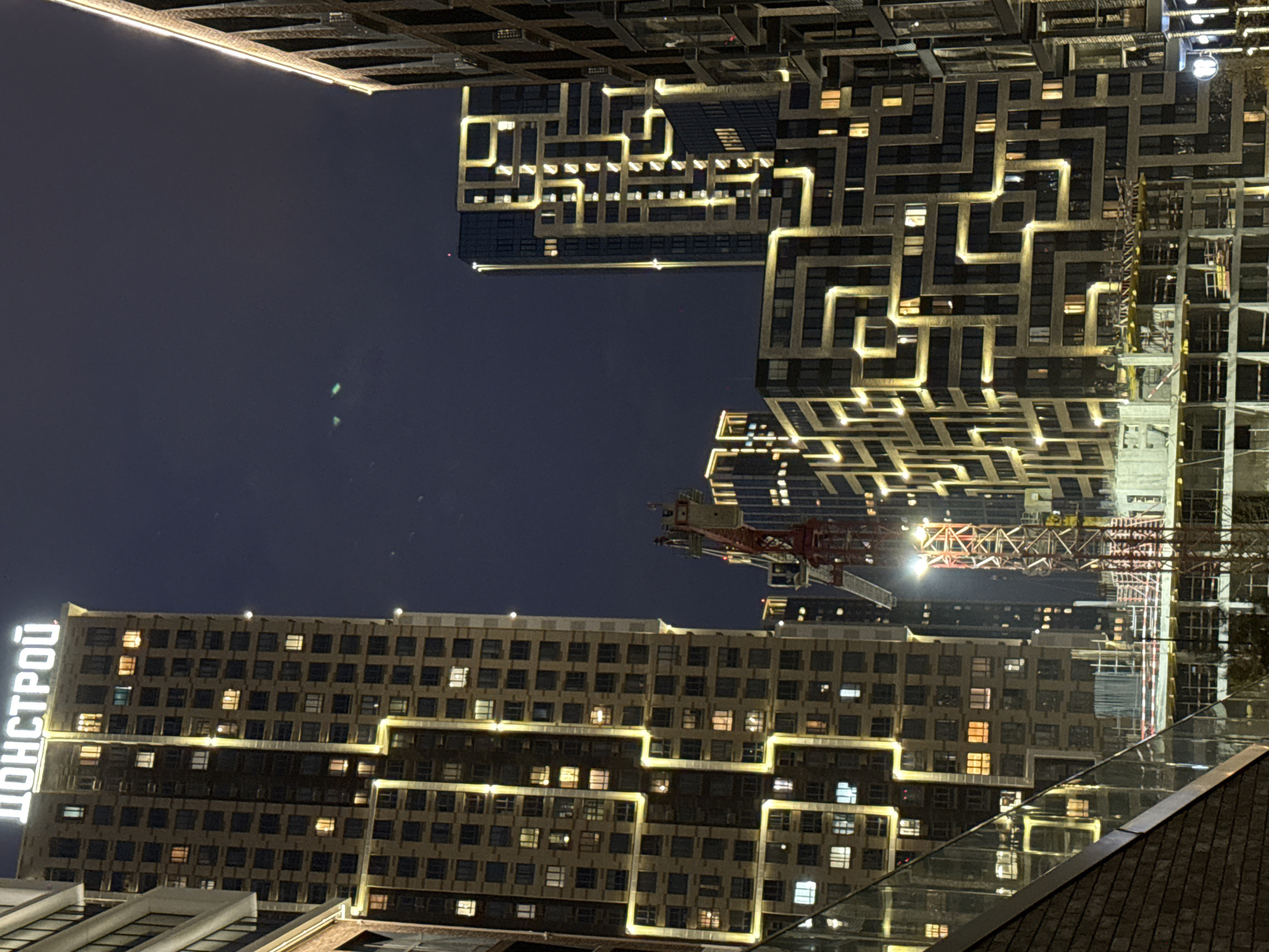 The image captures a night scene in Khimki, Russia, featuring several multi-story buildings illuminated with decorative lighting. The architecture is modern, with one prominent building displaying the name "ДОНСТРОЙ" in Cyrillic script on its facade. A construction crane is visible, suggesting ongoing building development. The environment appears urban, with evidence of street lighting and glass structures reflecting the surrounding illumination. The sky is dark, indicating it's likely nighttime.