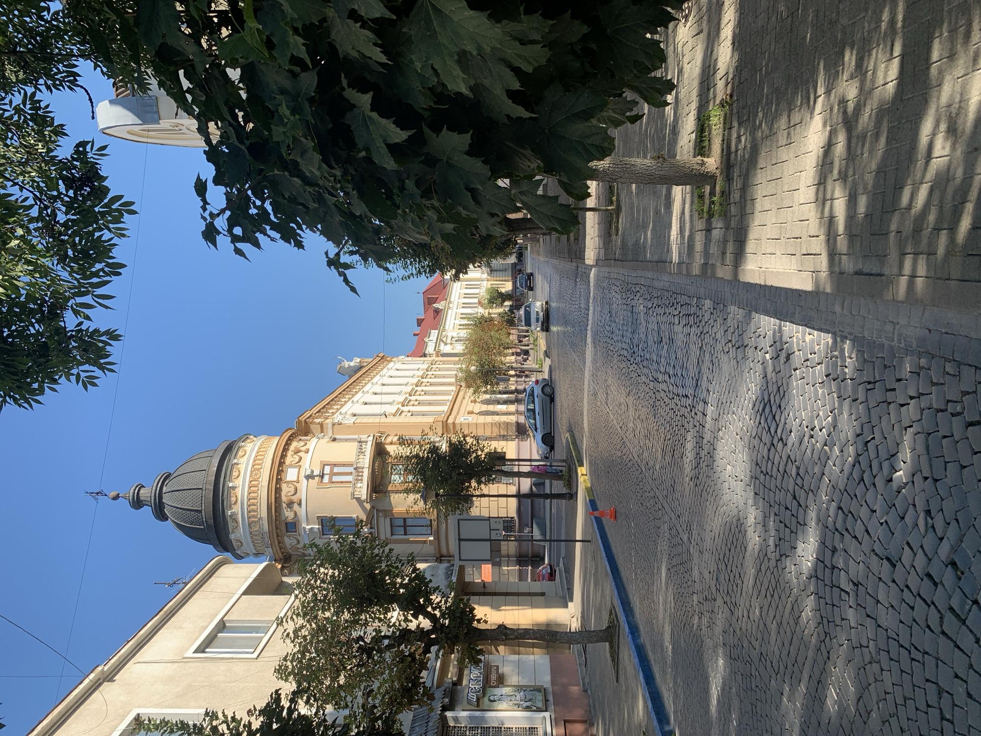 This image captures a sun-drenched cobblestone street in the city of Kolomyia, Ukraine. The street is lined with historic buildings, one of which features a prominent dome. The architecture suggests a European setting, with detailed facades and classical elements. Green trees with full canopies provide shade, casting dappled shadows on the pavement. Several cars are visible on the street, indicating it is a functional urban area. A lone orange traffic cone is placed on the side of the road. The sky is clear and blue, indicating fair weather. The overall impression is one of a quiet, picturesque European street scene on a bright day.