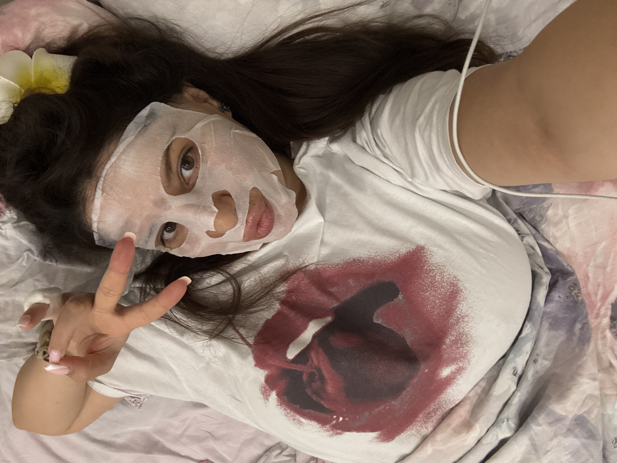 A woman wearing a white facial mask is lying on a bed covered in a floral patterned bedspread. She is wearing a white t-shirt with a dark red and black rose graphic on the front. She has long, dark hair with a white flower near her ear. Her left hand is raised in a peace sign, revealing manicured nails. A white wire, possibly a phone charger, is draped across her shoulder. The location is not discernible from the image and cannot be determined as Burgas, Bulgaria.