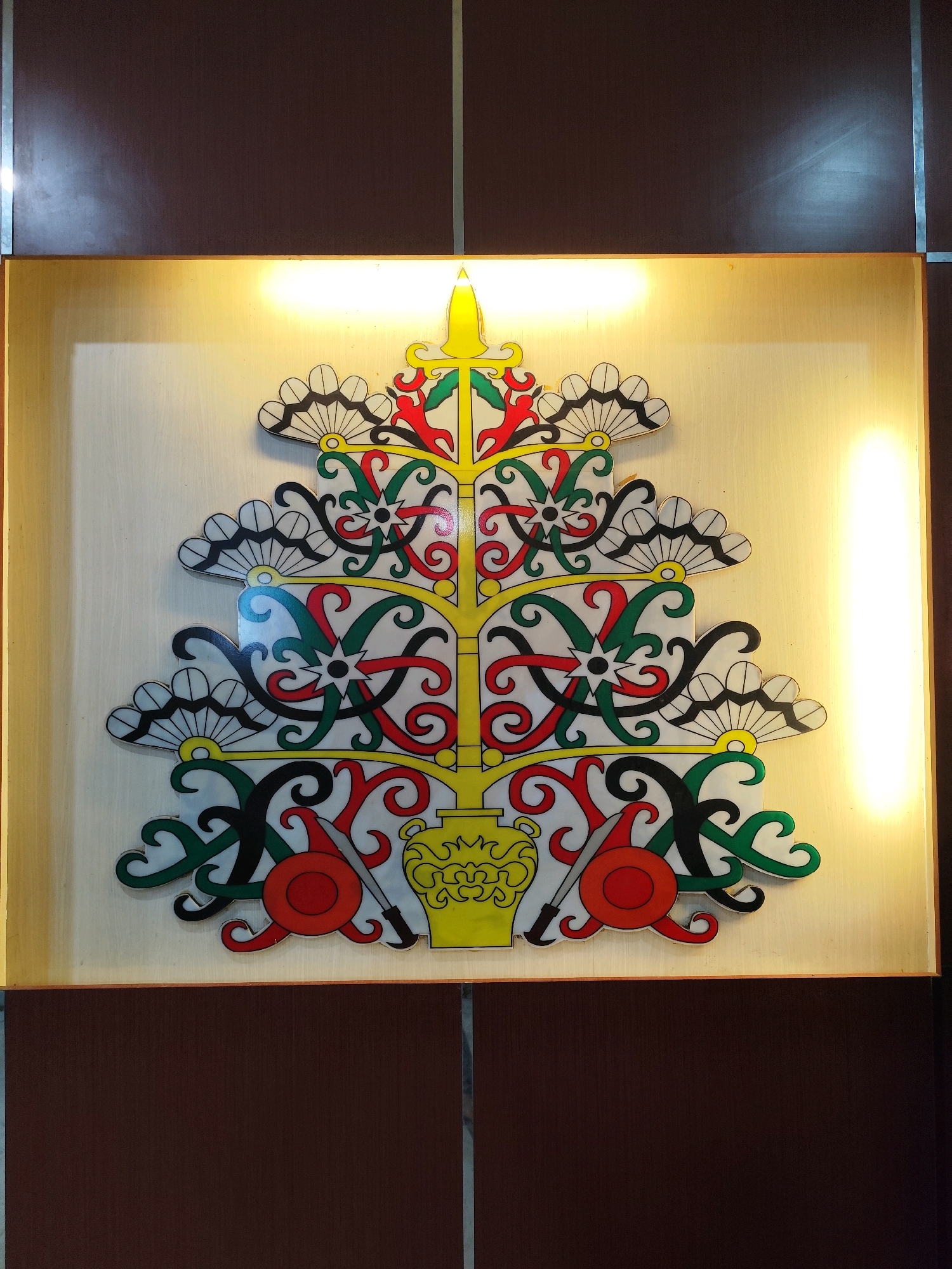 The image shows a decorative panel featuring a stylized tree-like motif with intricate patterns and vibrant colors, including red, green, yellow, black, and white. The motif appears to be a cultural emblem. The panel is mounted on a light-colored surface, possibly wood or laminate, and is illuminated by warm, diffused lighting from above, creating a glow around the edges. The overall presentation is framed by dark brown, textured panels on the top and bottom, with thin silver vertical dividers separating them. This decorative piece is a cultural emblem, likely representing the Dayak people of Borneo, and is a common sight in public buildings and spaces in Palangka Raya, the capital city of the Indonesian province of Central Kalimantan.