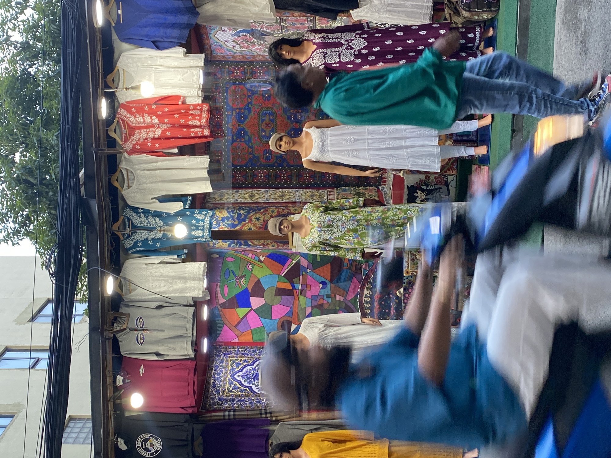 The image depicts a street scene in Kathmandu Metropolitan City, Nepal, featuring a storefront displaying a variety of textiles and clothing. In the foreground, a person on a blue scooter is captured in motion blur, wearing a teal shirt and white pants, suggesting movement through a busy area. Behind the scooter, a clothing stall is visible with mannequins dressed in traditional outfits. The stall showcases colorful fabrics and garments, including embroidered kurtis and dresses in shades of red, white, green, and purple. The background reveals more of the urban environment with a building and overhead wires.