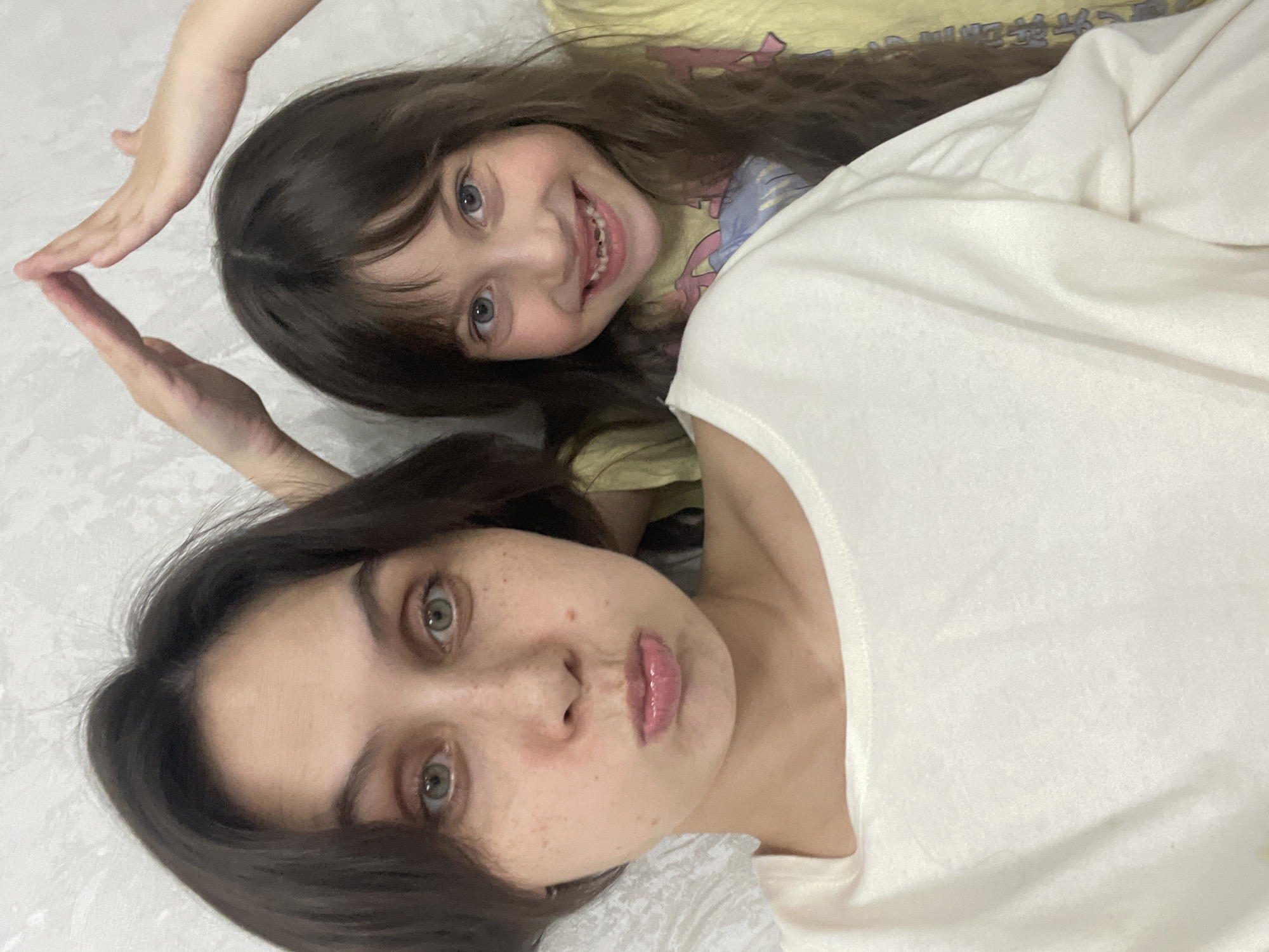 A candid, top-down image features two females, an adult and a child, lying close together on a light-colored surface, possibly a bed or floor, in an indoor setting. The image is rotated, showing them horizontally.
The adult, positioned on the left and closer to the viewer, has dark brown hair, light green eyes, and a light complexion with visible freckles. Her lips are slightly parted and appear glossy, and she wears a simple, light-colored (possibly white or off-white) top. Her expression is neutral or relaxed.
To her right, the child, with long dark brown hair and blue eyes, smiles widely, showing her teeth, indicating joy. She is wearing a light yellow top adorned with colorful patterns.
Above the child's head, two hands – one belonging to the adult and one to the child – are positioned with their fingertips touching, forming a V-shape or the outline of a heart. This gesture suggests a playful or affectionate interaction between the two subjects.
The background is a plain, bright, light-colored surface, providing a clean and uncluttered backdrop for the intimate moment. The overall mood of the image is warm and personal, capturing a sweet interaction between the individuals. There is no visible text.