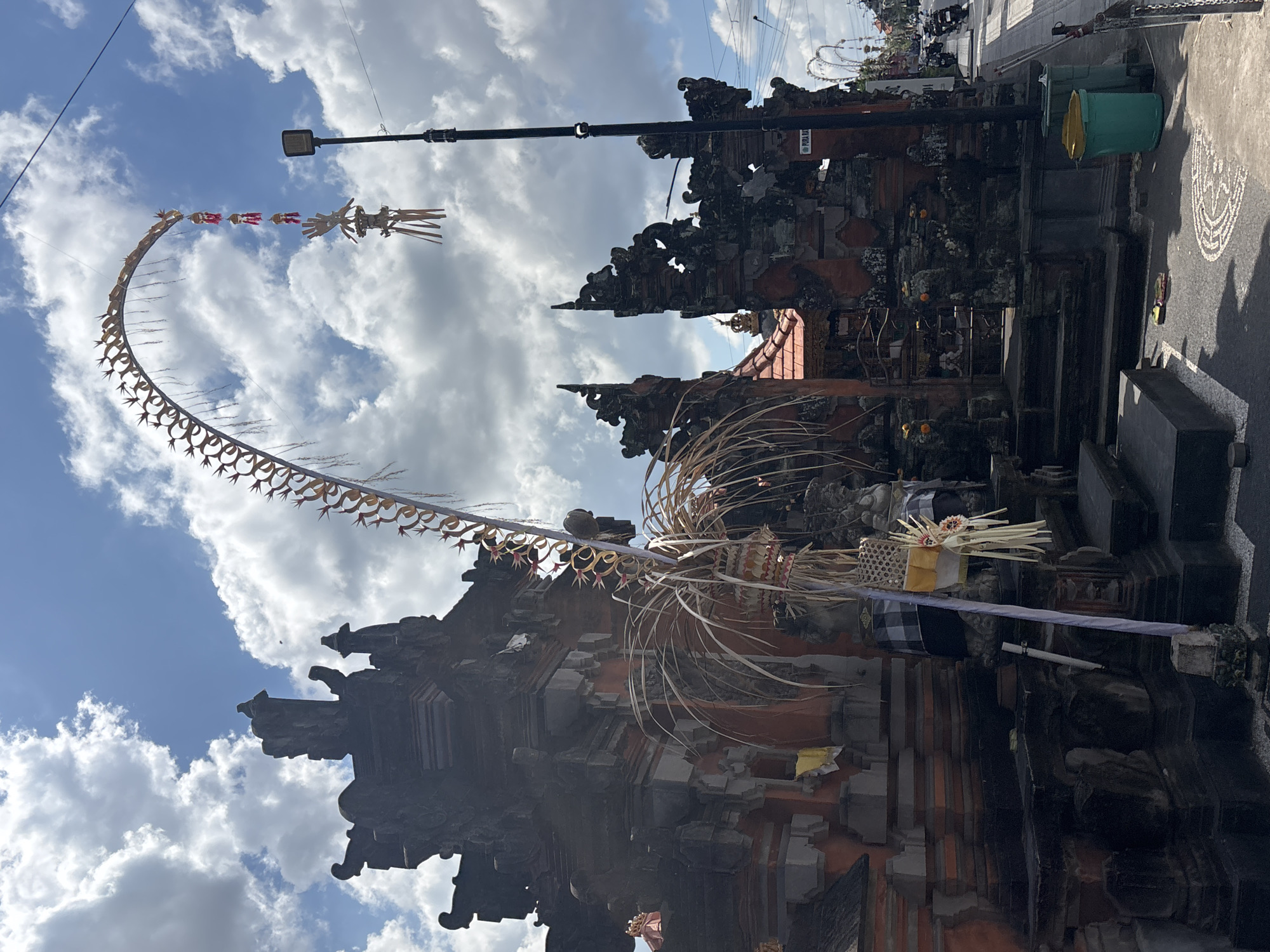 A Balinese temple gate, known as a Candi Bentar, is visible on the left side of the image. The gate is intricately carved from dark volcanic stone and reddish-orange brick, with tiered roofs and ornate sculptures. A large, curved ceremonial bamboo structure, adorned with small red and gold decorations and straw ornaments, arches over the street from the temple. The sky above is a bright blue with scattered white clouds. A modern street light stands on the right side of the image, near the curb. The street extends to the right, with several motorcycles parked or riding in the distance. Two green and yellow trash cans are placed near the sidewalk. The ground in front of the temple features a circular pattern made of white pebbles.