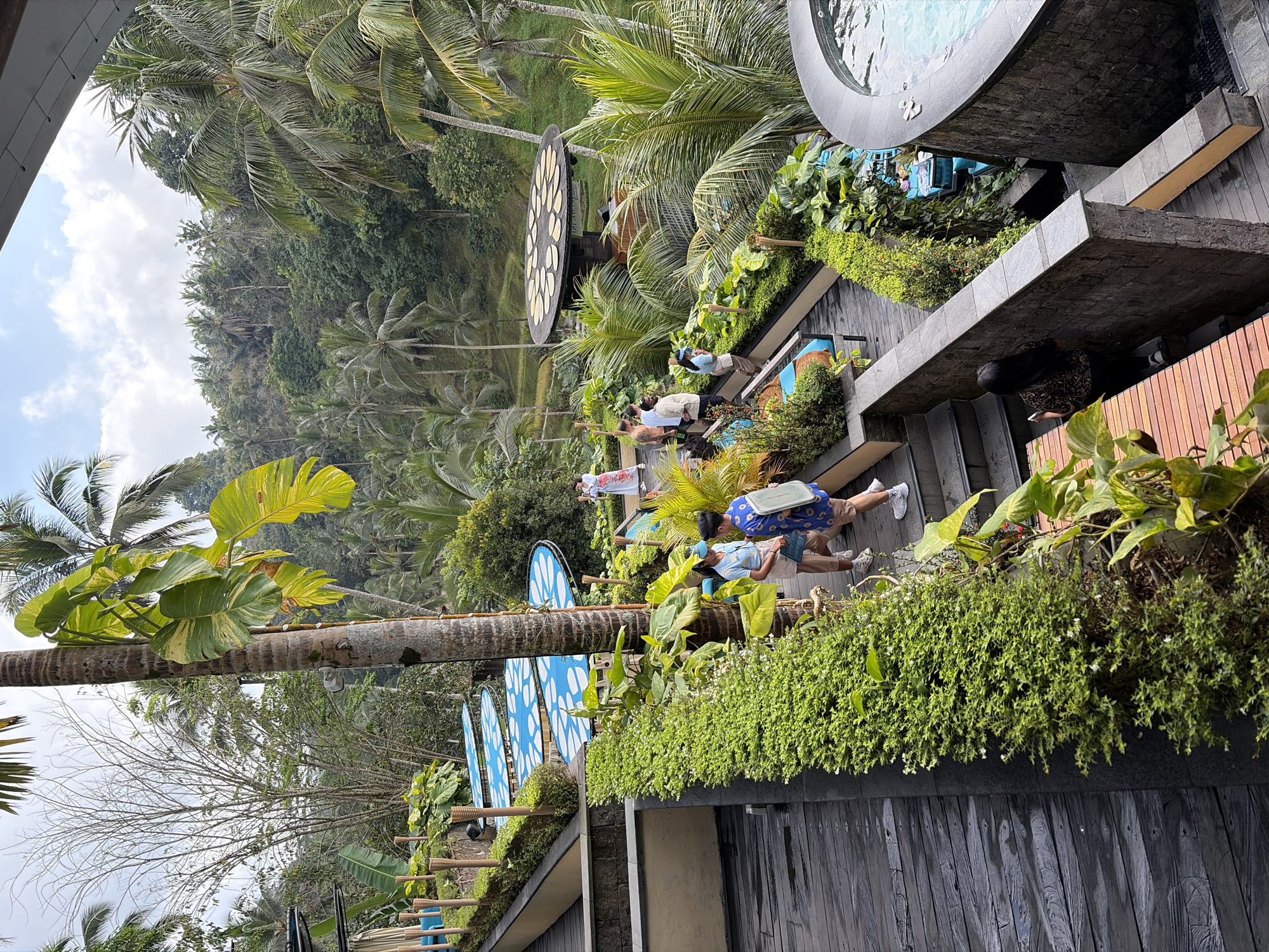 The image depicts a scenic overlook in Tegallalang, Indonesia, characterized by lush tropical vegetation and a view of a valley filled with palm trees. In the foreground, a wooden walkway lined with meticulously maintained green hedges leads the eye down a series of stone steps. Several people are visible. Two individuals, a man and a woman in casual attire, are engaged in conversation on the walkway. Further down, a group of people, including at least two women and one man, are seen walking. In the middle ground, a circular structure with a thatched roof, possibly a gazebo or pavilion, is adorned with flat, round objects arranged in a circular pattern. Several blue-tiled seating areas with white accents are scattered throughout the scene, hinting at a resort or recreational area. In the background, a dense jungle landscape dominates, with terraced rice paddies visible in the distance. The sky is partly cloudy, suggesting a pleasant daytime. A glimpse of a circular hot tub or pool is visible on the right side of the frame, adding to the impression of a luxurious getaway.