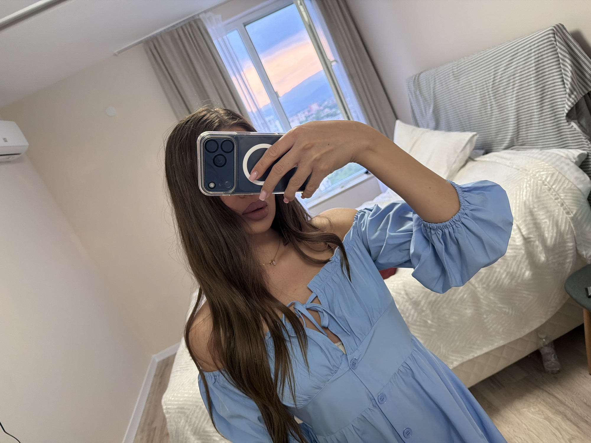 The image captures a person taking a reflection selfie in a room located in Plovdiv, Bulgaria. The central subject, whose face is predominantly obscured by a dark-colored smartphone held in their right hand, exhibits long, dark brown hair and is wearing a light blue, off-the-shoulder dress with gathered sleeves and front buttons. A delicate gold necklace is visible around their neck, and pink lipstick is apparent on their lower face. The smartphone, encased in a transparent cover, clearly displays a MagSafe ring and a multi-lens camera array.
The room features light beige walls, a light wood laminate floor, and a large bed with white bedding, including multiple pillows. A grey and white striped fabric is draped over the bed's headboard. A white wall-mounted air conditioning unit is affixed to the left wall. In the background, a large window, framed by light grey curtains, reveals an outdoor scene characteristic of Plovdiv: an urban area with buildings spreading towards distant hills or mountains, all set against a sky painted with hues of blue, pink, and orange, indicative of either sunrise or sunset. A small, dark round table with a wooden leg is partially visible on the right side of the bed. A small, circular light fixture is visible on the white ceiling.