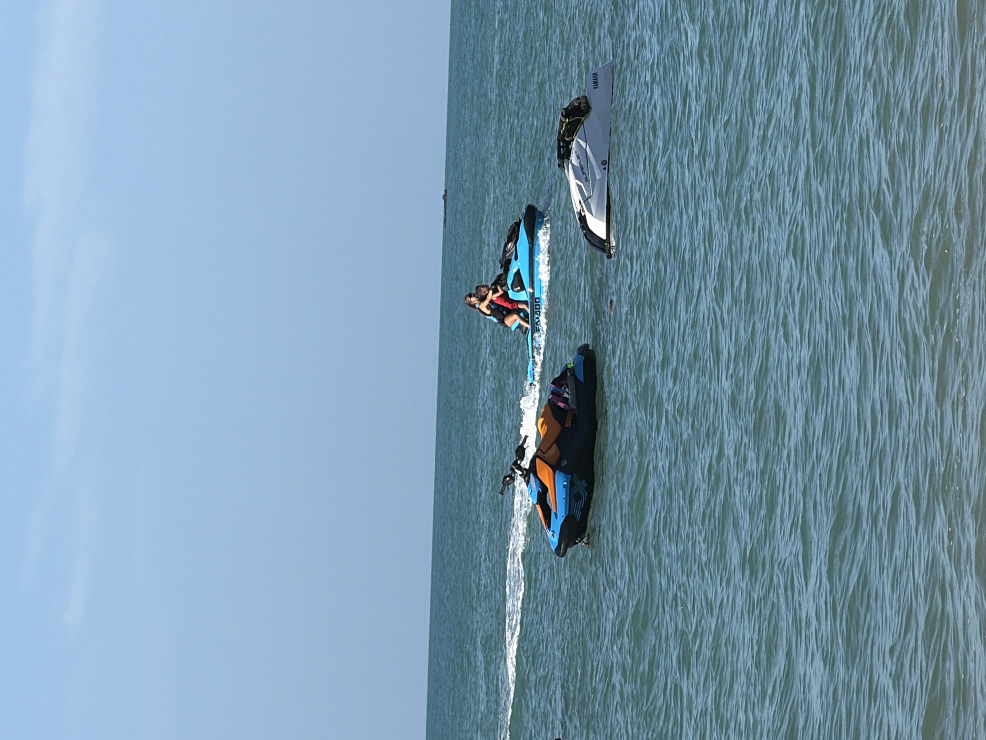 This image captures a bright, clear day at sea, likely off the coast of Ko Samui District, Thailand, given the calm, turquoise waters and blue sky. The scene features several jet skis on the water. In the foreground, a blue and orange jet ski is stationary. To its right and slightly further back, a blue jet ski is in motion, carrying two people. Further to the right and in the foreground is a white jet ski, also stationary. The water is slightly choppy, with white wakes visible behind the moving jet ski. The horizon is visible in the distance, separating the sea from the clear sky.