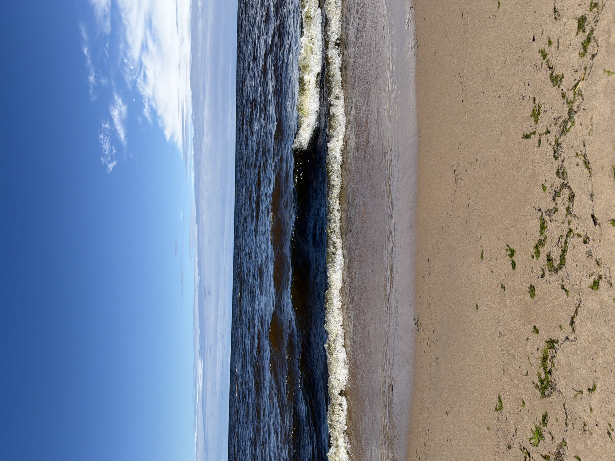 This is a vertical, eye-level shot of a sandy beach in Lilaste, Latvia. The image is split into three horizontal sections: sky, sea, and beach. The sky is a vibrant blue with scattered white clouds, particularly on the right side, some of which are thin and wispy. The sea is a deep, dark blue, with small to medium-sized waves breaking on the shore. The waves have white foam crests. The beach is composed of light brown sand, with some scattered patches of green seaweed. The water meets the sand in a gentle slope, with the wet sand appearing darker and more reflective. No people or man-made objects are visible in the frame.