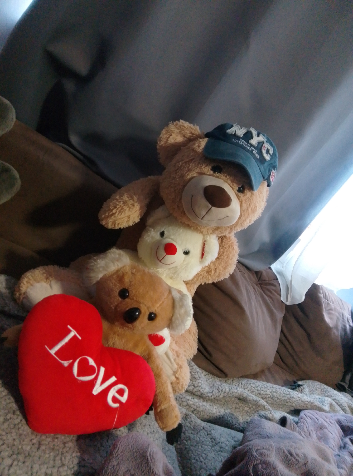 The image depicts a close-up of three teddy bears and a red heart-shaped pillow. A large, light brown teddy bear is in the foreground, wearing a blue baseball cap that says "NYC". It is holding a smaller, white teddy bear, which in turn is holding another, smaller light brown teddy bear. The red heart pillow, with the word "Love" written in white script, is placed in front of the bears. The bears and pillow are situated on a soft, textured surface that appears to be a bed or couch covered with blankets. In the background, brown and gray curtains are visible, suggesting an indoor setting. There is no indication of the city of Wedel, Germany, in the image.