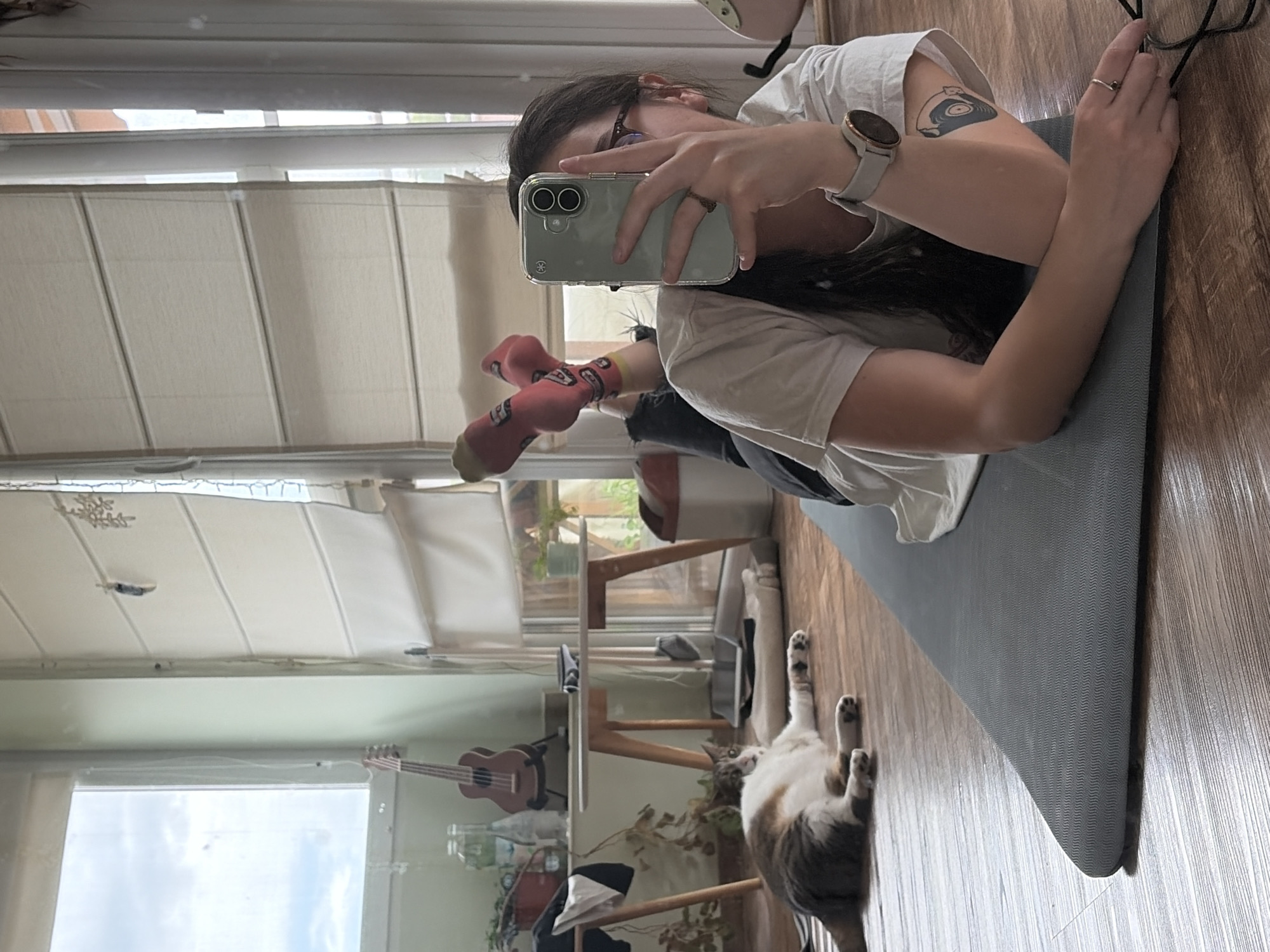 The image captures a casual indoor scene featuring a person and a cat. The primary subject is a person, appearing to be female, with long dark hair and glasses. She is lying on her stomach on a grey yoga mat, propped up on her elbows, and is taking a mirror selfie with a light green smartphone. She wears a white t-shirt, a smartwatch on her left wrist, and a ring on her right hand. A tattoo of a stylized vinyl record is visible on her left arm. Her feet, clad in red patterned socks, are also visible.
Beside the yoga mat, a calico cat with white, brown, and black fur lies on its back, stretching its paws in a relaxed posture.
The setting is an indoor room, likely a living area or bedroom, characterized by large windows letting in natural light. Cream-colored blinds cover the upper part of the windows. A light wooden floor extends throughout the visible area. In the background, a ukulele rests on a stand next to a window, and a wooden table holds potted plants and a water bottle, suggesting a cozy, lived-in environment.
The activity taking place is a relaxed moment, with the person capturing a selfie while lounging on a yoga mat, and the cat resting comfortably nearby. The natural light indicates it is daytime, with an overcast sky visible through the windows. No identifiable text is visible.