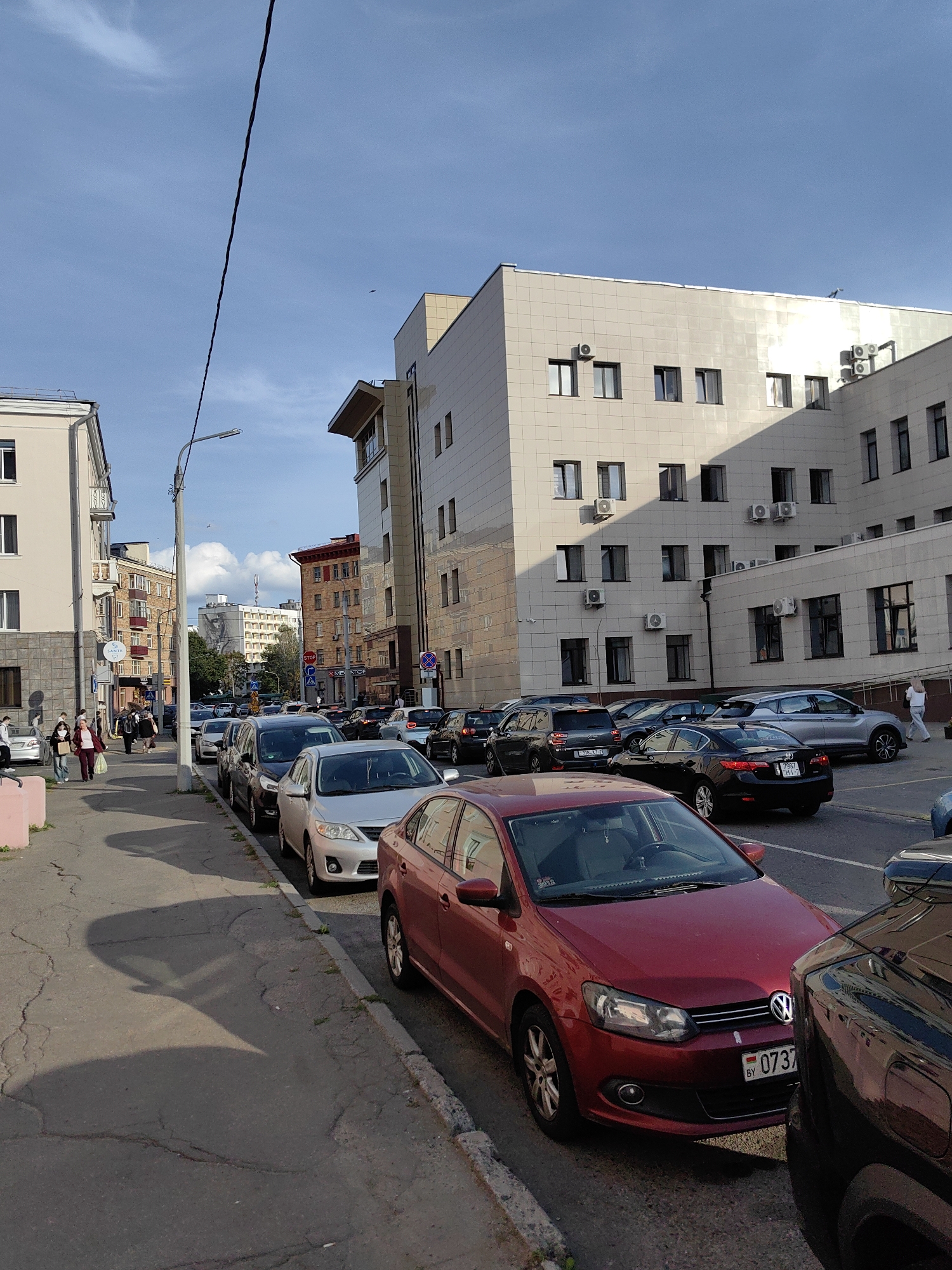 This is a medium-angle, eye-level, full shot of a street in Minsk, Belarus, on a sunny day. The scene captures a row of parked cars lining the side of the road, with a sidewalk and buildings on the left and right.
The foreground is dominated by the front of a red Volkswagen Polo, parked on the street. To its right, partially visible, is the front fender of a dark-colored SUV. The street itself is asphalt, with visible cracks and a distinct curb.
A sidewalk runs parallel to the street on the left, featuring a light pink painted curb. Several people are walking on the sidewalk, some with shopping bags, and some appear to be wearing masks. Buildings flank the street, with the most prominent on the right being a modern, light-colored, multi-story building with numerous windows and air conditioning units. On the left, a lighter colored, older building with balconies and windows is visible.
In the background, the street continues, lined with more parked cars and buildings of varying architectural styles and colors. The sky above is a clear blue with a few white clouds and a thin, dark wire stretching diagonally across the frame. The overall impression is of a typical urban street scene.