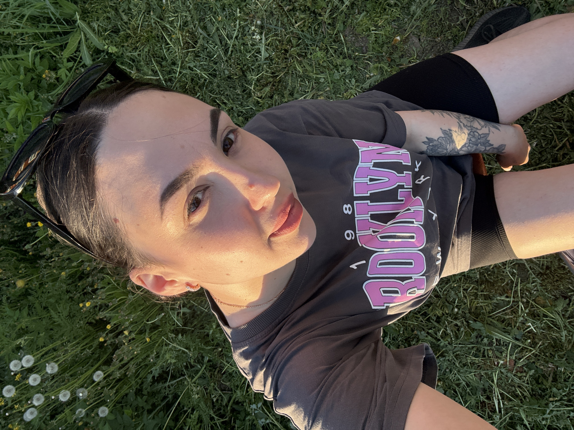A woman is lying on a grassy surface. The woman is fair-skinned and has dark hair pulled back, with sunglasses propped on her head. She is wearing a grey t-shirt with the word "BROOKLYN" in large pink letters, and black shorts. The t-shirt has the number "98" on it as well. A tattoo is visible on her right arm. The grass beneath her is dotted with dandelions. Part of a black shoe is visible in the upper right corner. The woman is looking directly at the camera.
The city of Фоки, Russia is not relevant to the context of this image.