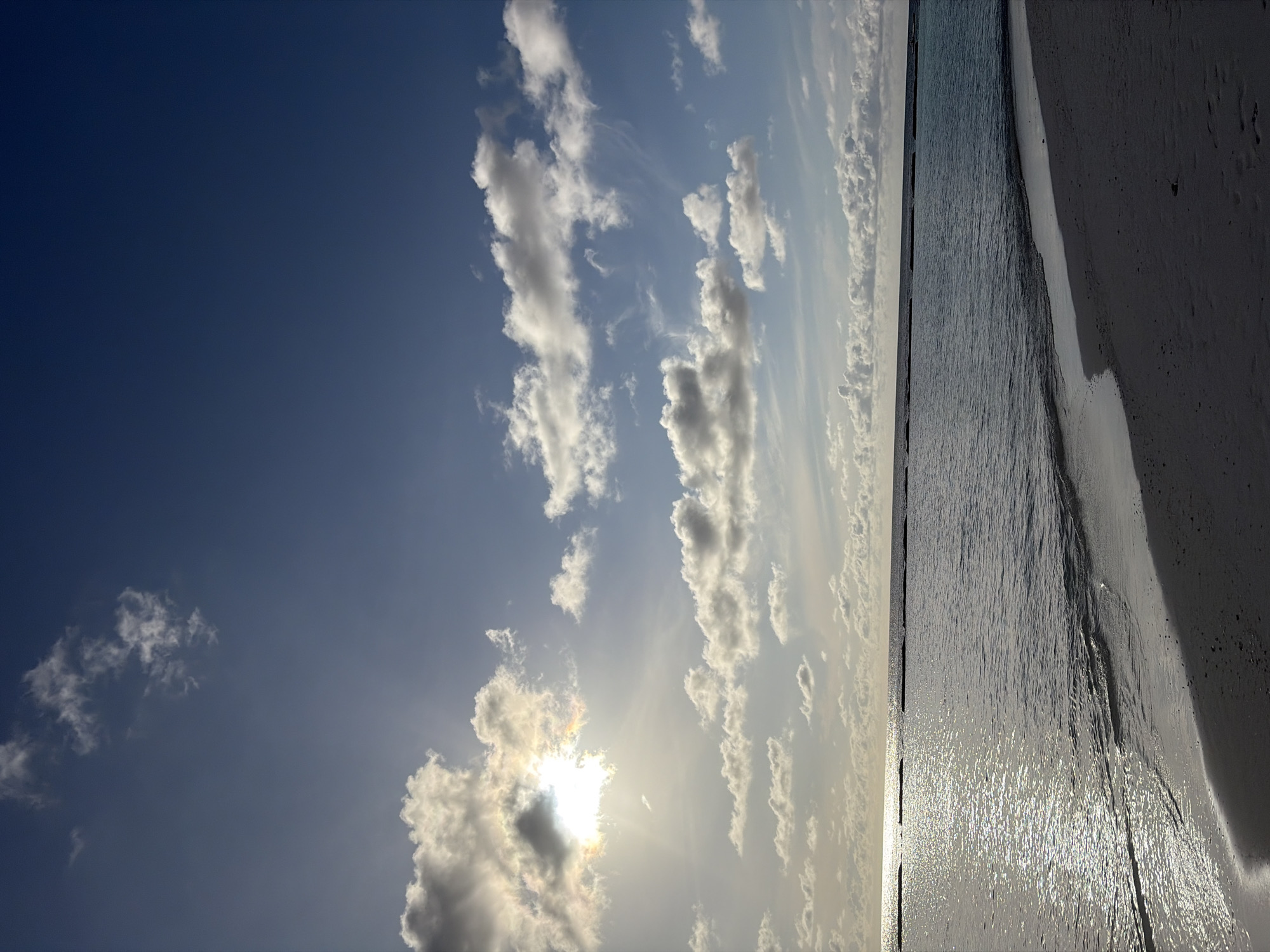 This is a photograph of the beach and ocean in Halaveli, Maldives. The image features a sandy shore in the foreground, meeting the calm, dark blue water of the ocean. The sun is visible in the upper left quadrant of the image, partially obscured by bright white and grey clouds, casting a shimmering reflection on the water's surface. The horizon line is clearly defined, separating the sea from a pale blue sky dotted with scattered white clouds. There are no people or animals visible in the frame. The scene depicts a tranquil beach environment.