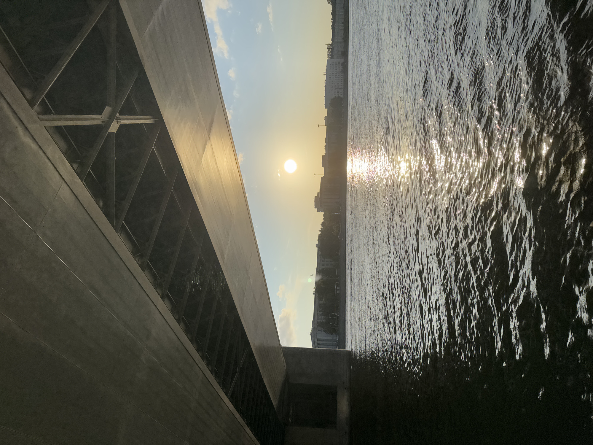 An under-bridge view of a river in Saint Petersburg, Russia, with the sun setting over the city skyline. The underside of the bridge, made of concrete and steel, is visible in the foreground. The river is calm with gentle ripples reflecting the sunlight. In the distance, buildings line the riverbank, and the setting sun casts a golden glow on the water and the sky. The sky is partly cloudy with a few white clouds scattered across the blue expanse. No people or specific actions are visible. The scene depicts a serene urban landscape during sunset.