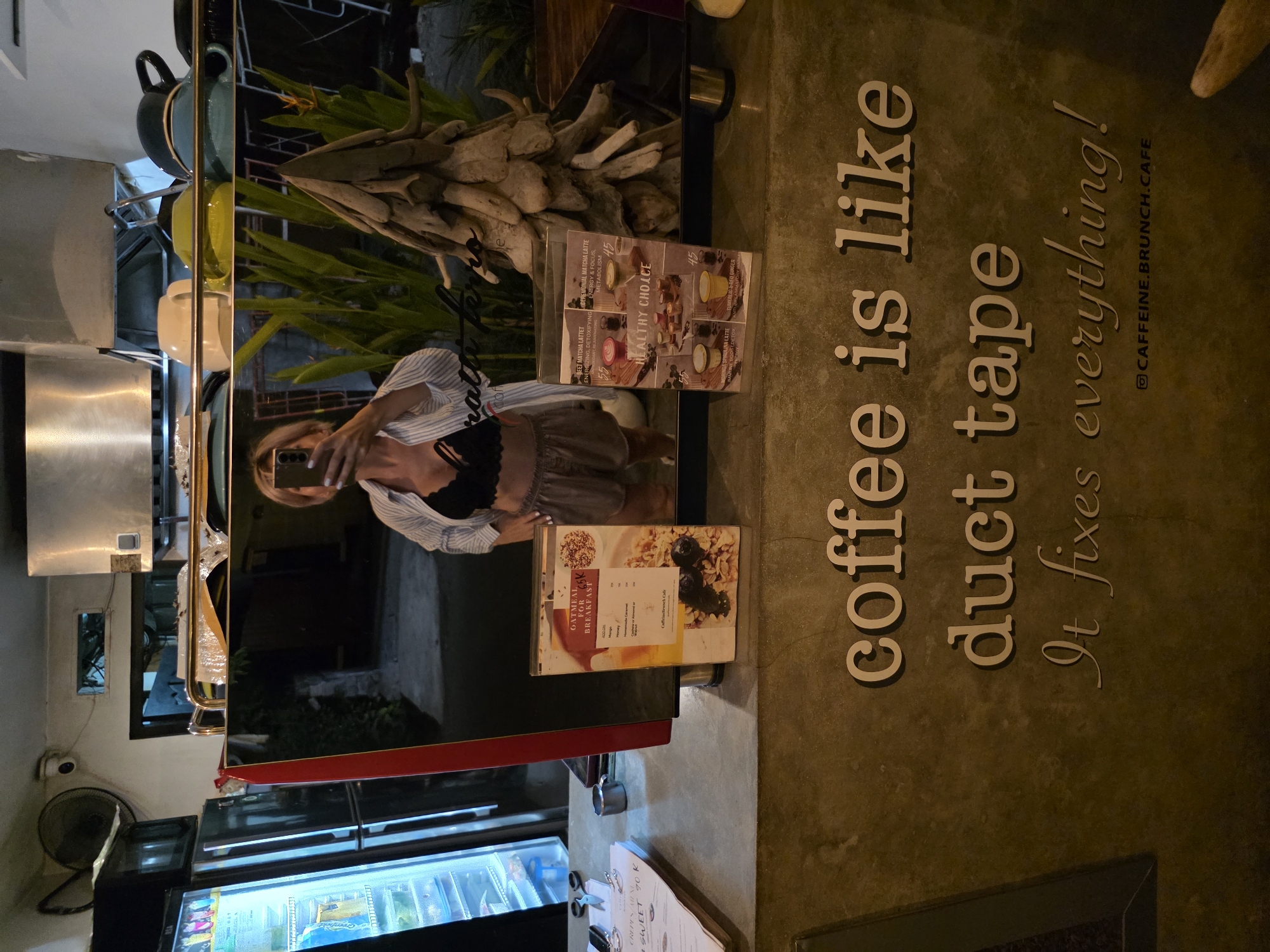 A full-body, vertical mirror selfie is taken in what appears to be a cafe or restaurant. The person in the photo, wearing a striped shirt and shorts, is holding up a smartphone to capture the reflection. The cafe's interior is visible in the background, including a display case with food items, plants, and decorative driftwood. The floor has large, raised text that reads "coffee is like duct tape it fixes everything!" and a social media handle "@CAFFEINE.BRUNCH.CAFE". Menus are displayed on a glass surface, showing items like "Oatmeal for Breakfast" and "Beet Matcha Latte". The lighting suggests it could be evening or the cafe is dimly lit. The scene appears to be in North Kuta, Indonesia.