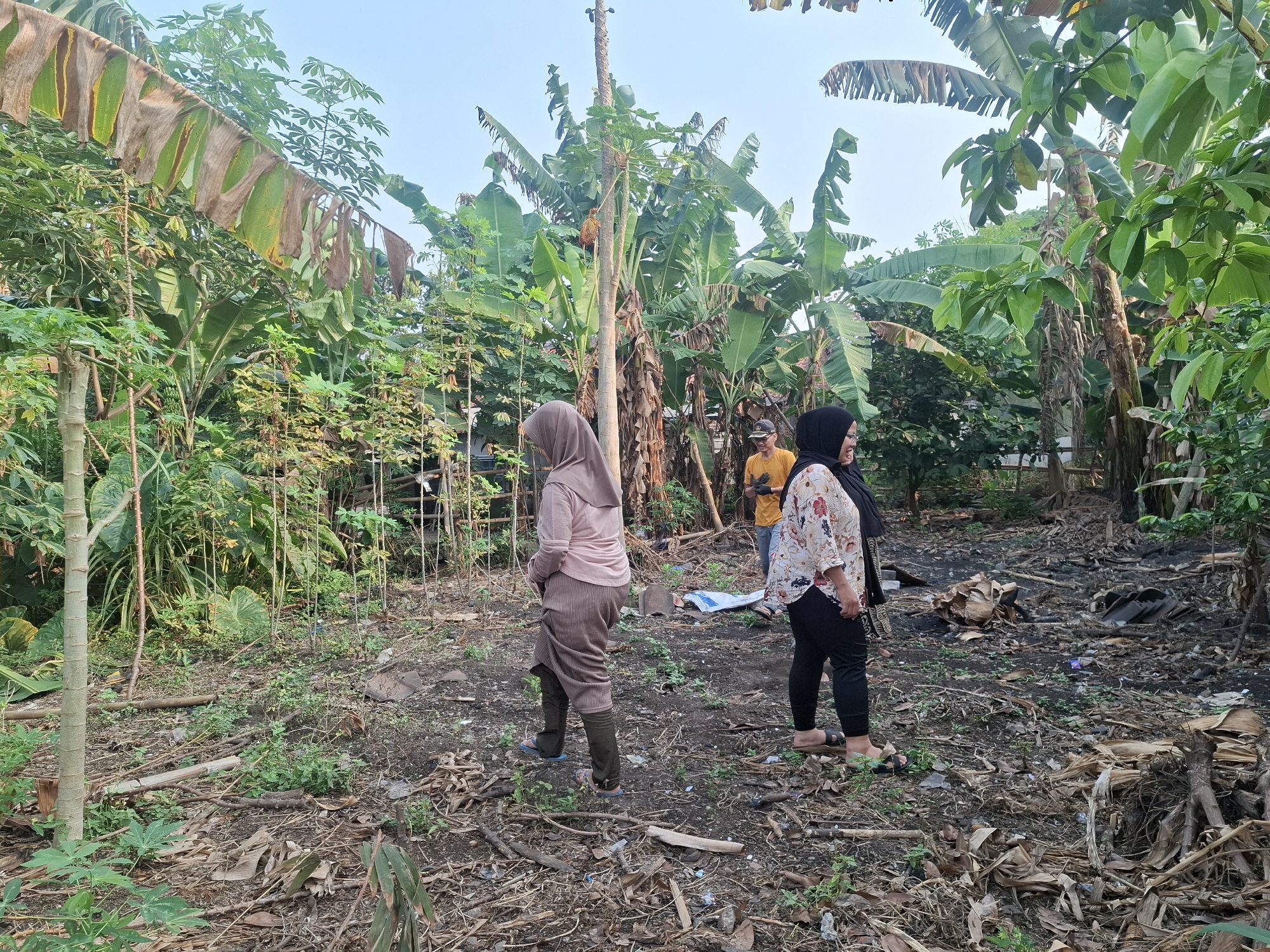 In Bojong, Indonesia, two women in modest Islamic attire stand in a cleared area surrounded by dense tropical vegetation. The woman on the left wears a pale pink hijab and a long, striped brown skirt over olive-green leggings, with blue sandals. She is facing away from the camera, looking towards the left. The woman on the right wears a black hijab and a white floral print blouse with black leggings and black sandals. She is facing towards the left, partially obscuring the man standing behind her. The man, visible in the center background, is wearing a bright yellow t-shirt and dark pants. The ground is covered in dry leaves, branches, and some debris, suggesting an outdoor, possibly agricultural or undeveloped setting. The background is filled with lush banana trees and other foliage, indicating a warm, humid climate. The overall impression is a candid moment captured in a rural or semi-rural Indonesian landscape.