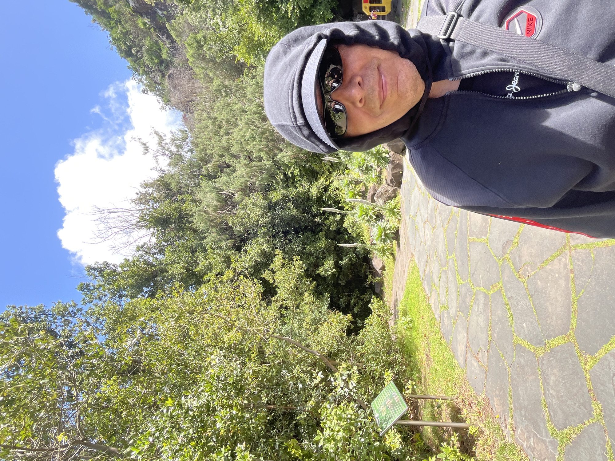 A selfie taken at an outdoor location with dense green foliage and a blue sky with scattered clouds. The person in the foreground is wearing a dark hoodie and sunglasses, with a shoulder bag. The background shows a paved pathway and a sign with green text in front of a lush landscape. The location is Las Palmas, Spain.