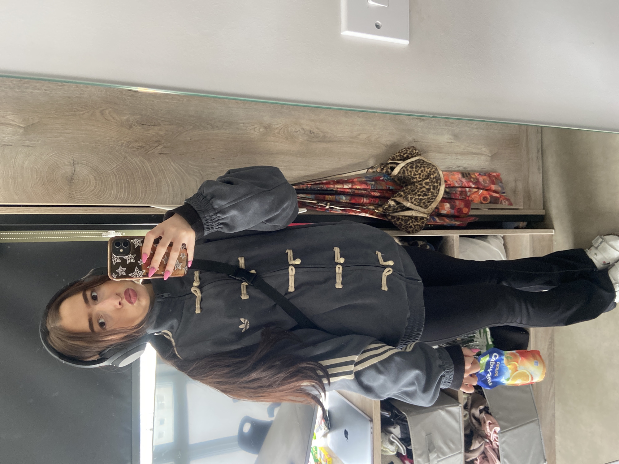 A woman is taking a selfie in a mirror. She is wearing headphones, a grey hooded Adidas jacket, and black pants. She has long brown hair. She is holding a phone in her left hand, a Capri Sun in her right hand. The background shows a room with a wooden shelf, a laptop, storage bins, and a light switch on the wall. The location is not identified as London, United Kingdom, based on the provided image.