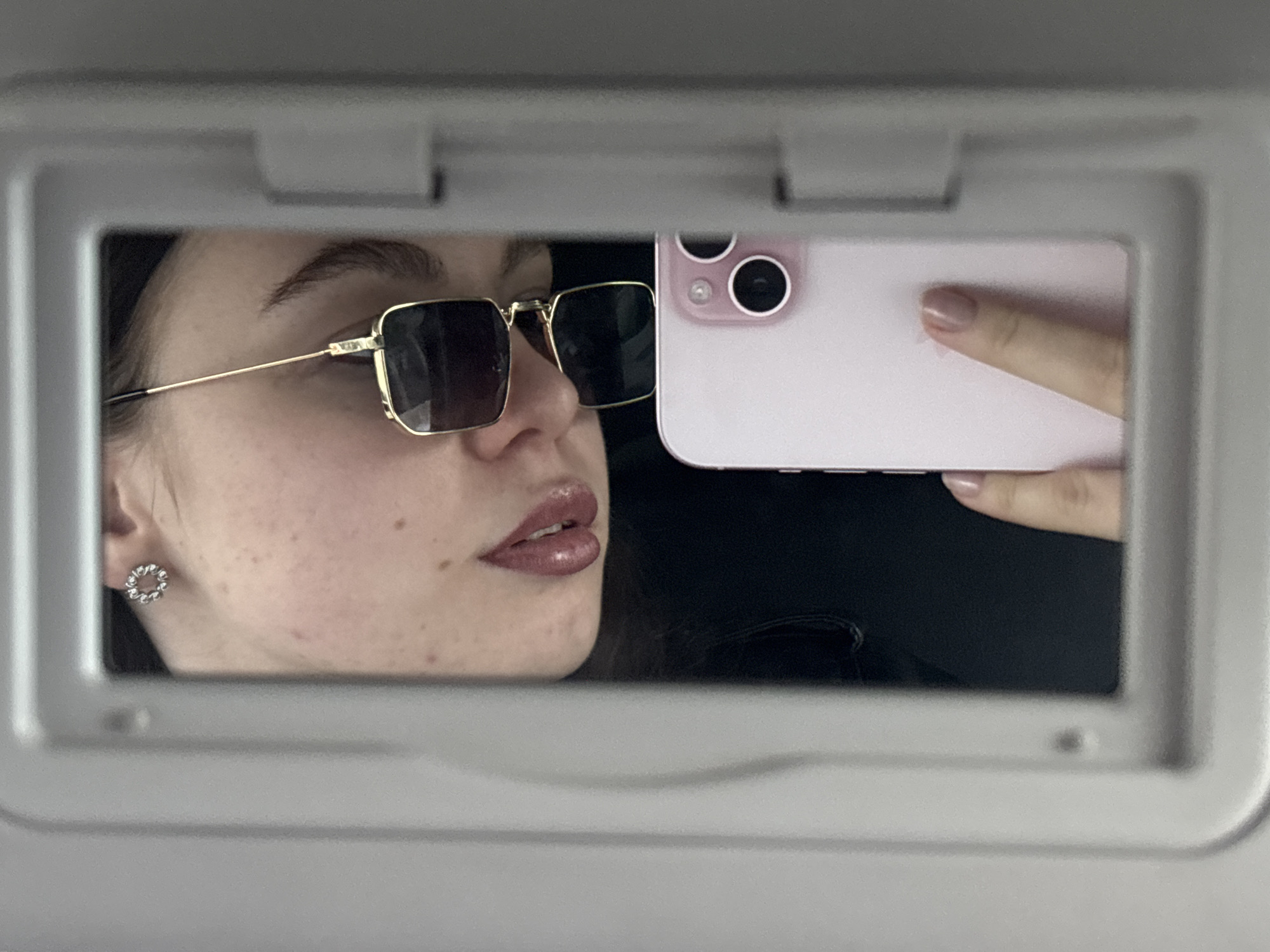 A woman is visible in a selfie taken in a car's visor mirror. She's wearing gold-framed sunglasses, and a floral earring is visible. A pink smartphone is in the frame, partially obscuring the reflection. The backdrop is black, possibly the car's interior. The location is unlikely to be Perm, Russia, as there is no context in the image to associate it with the city.