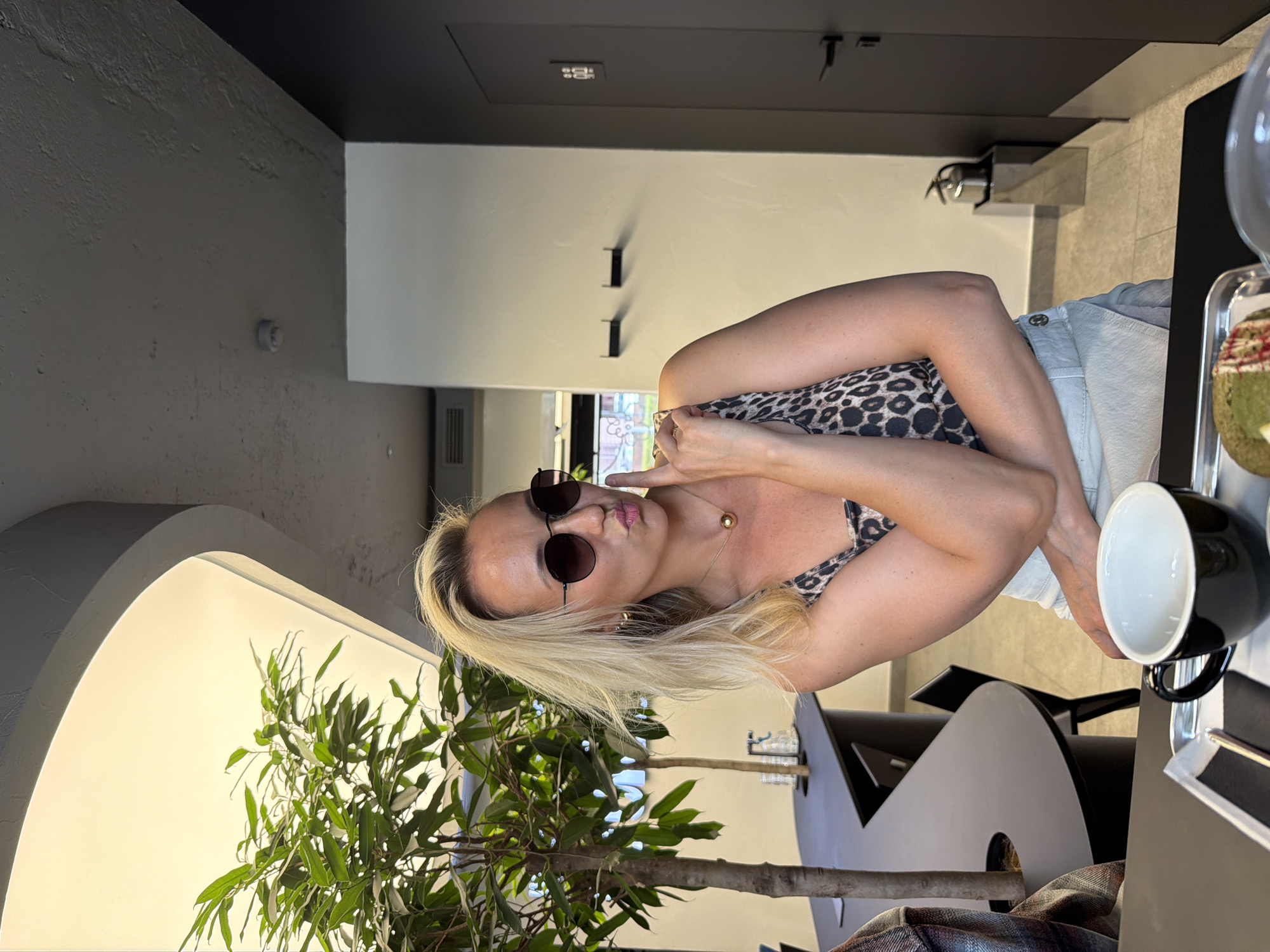 A woman with blonde hair is sitting at a table in a cafe. She is wearing sunglasses, a leopard print top, and light-colored shorts. She is posing for the camera, with one hand near her face and her lips pursed. On the table in front of her is a black mug, a dessert on a tray, and a plant. The cafe appears to be modern with dark walls and neutral-colored furniture. The lighting suggests it could be daytime. The woman appears relaxed and playful. There is no visible text in the image.