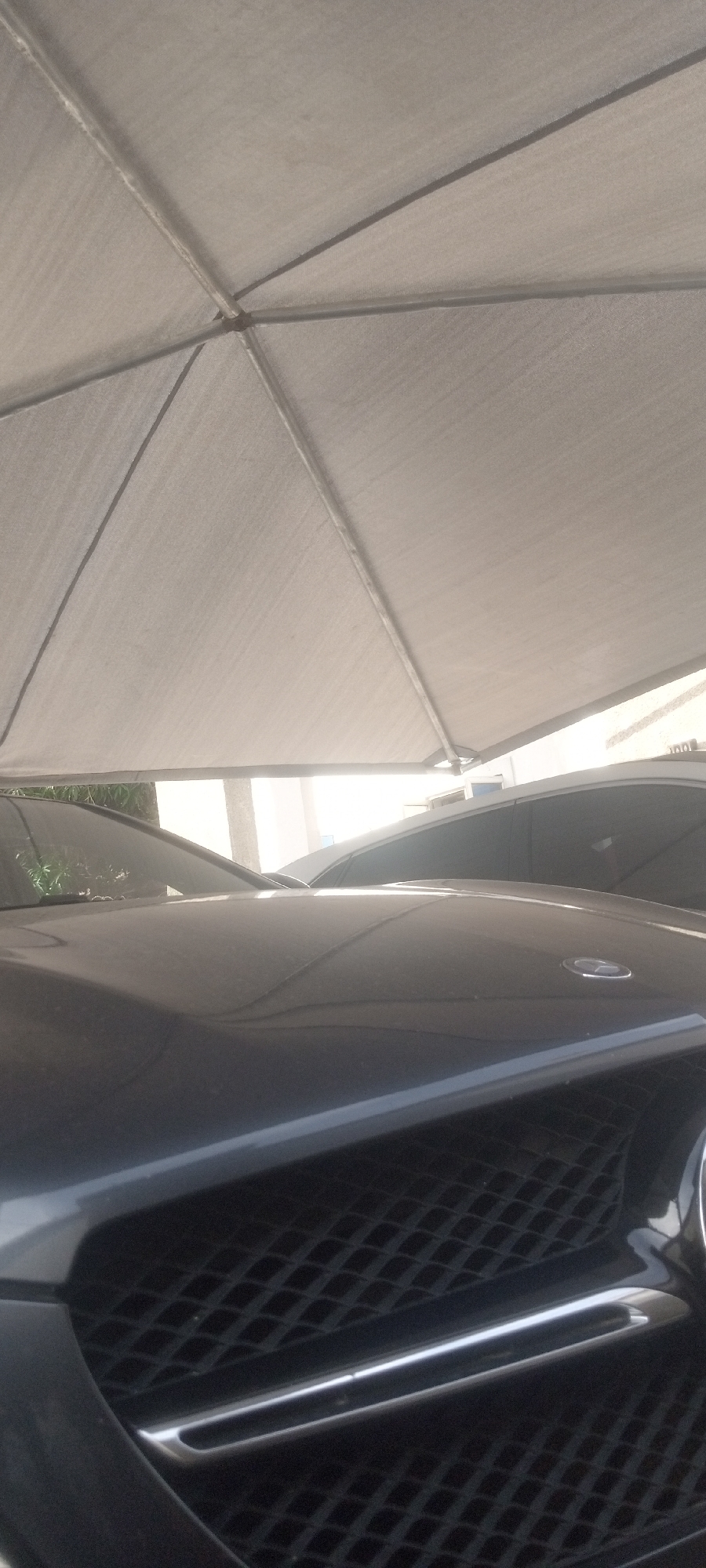 The image shows the front of a black Mercedes-Benz vehicle, specifically focusing on the grill and hood, parked under a gray awning. The location is most likely Narayi, Nigeria, though this cannot be confirmed solely from the visual data. The area seems to be an open-air parking or storage space, with at least one other vehicle visible in the background. There are no people visible in the image. The details provide no indication of specific actions or interactions taking place.