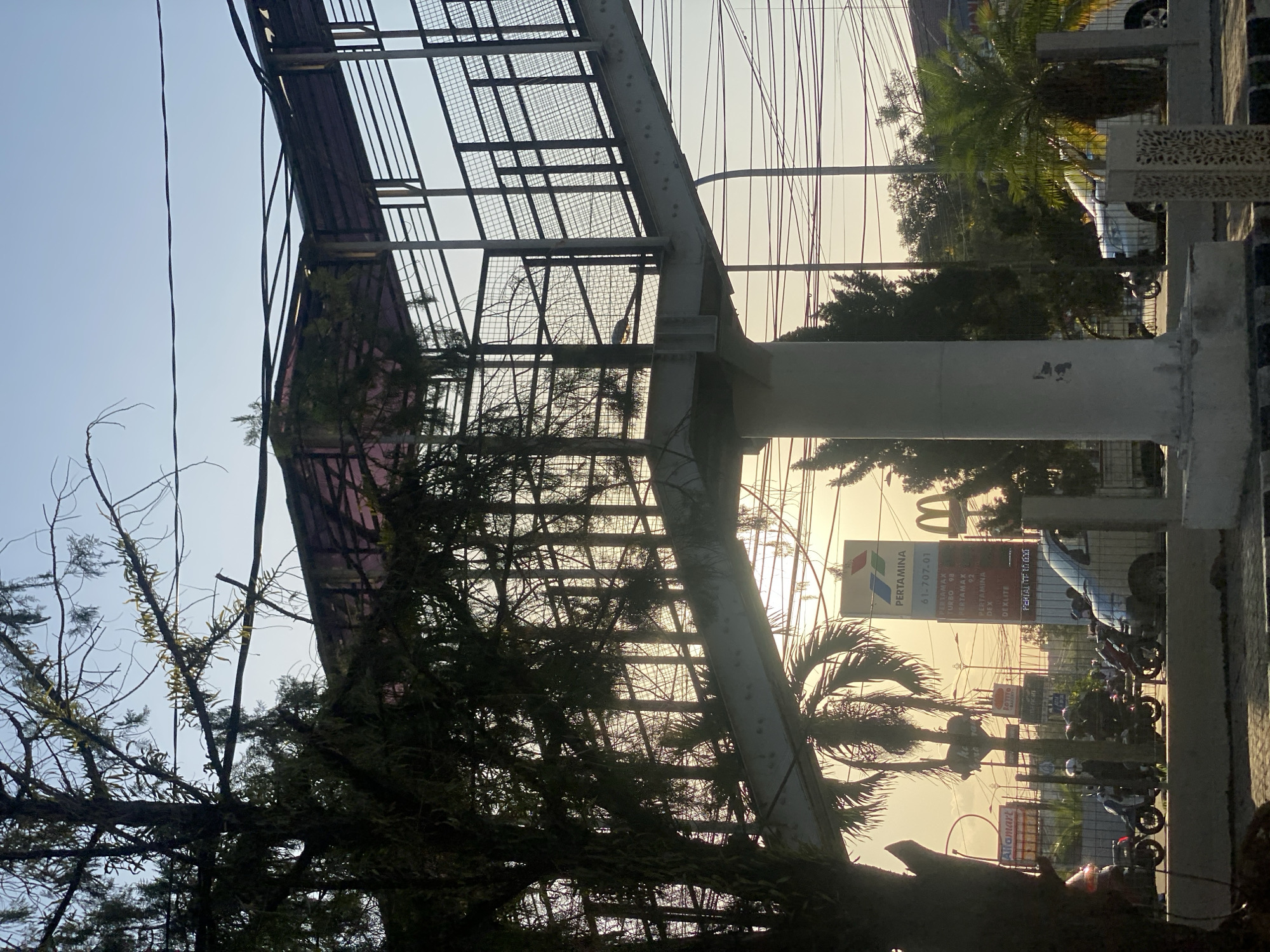 The image shows an elevated pedestrian walkway with a metal frame and a red translucent roof, supported by a concrete pillar. The walkway is surrounded by trees, including a large, dark-leafed tree in the foreground and palm trees in the background. In the distance, various buildings and signs are visible, including a Pertamina gas station sign with fuel prices listed, a McDonald's sign, and an Alfamart sign. Several motorcycles are parked or being ridden on the road below the walkway. The scene is illuminated by the warm light of a sunset, casting long shadows and creating a hazy atmosphere. The location is identified as Banjarbaru, Indonesia.