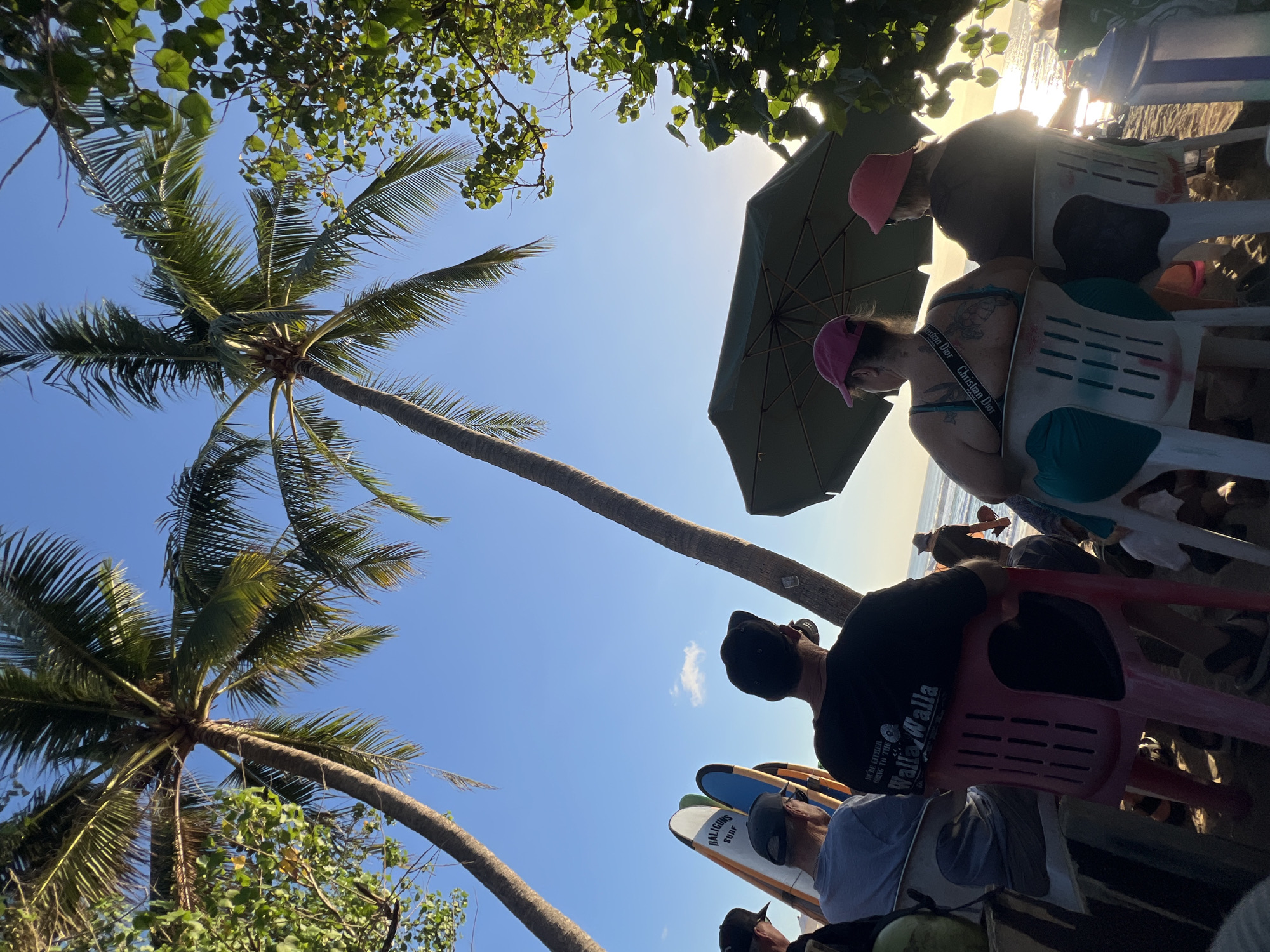 The image depicts a beach scene in Kuta, Indonesia, likely during sunset. In the foreground, several people are seated on chairs facing the ocean, some under a large green umbrella. Among them, a person wearing a black t-shirt with text that reads "WE'RE EITHER GOING TO THE WALLA WALLA" is visible. To the left, a collection of surfboards leans against a structure, and two palm trees frame the sky. The ocean is visible in the background, with waves and the setting sun casting a warm glow on the water. The overall atmosphere suggests a relaxed, casual gathering on the beach.