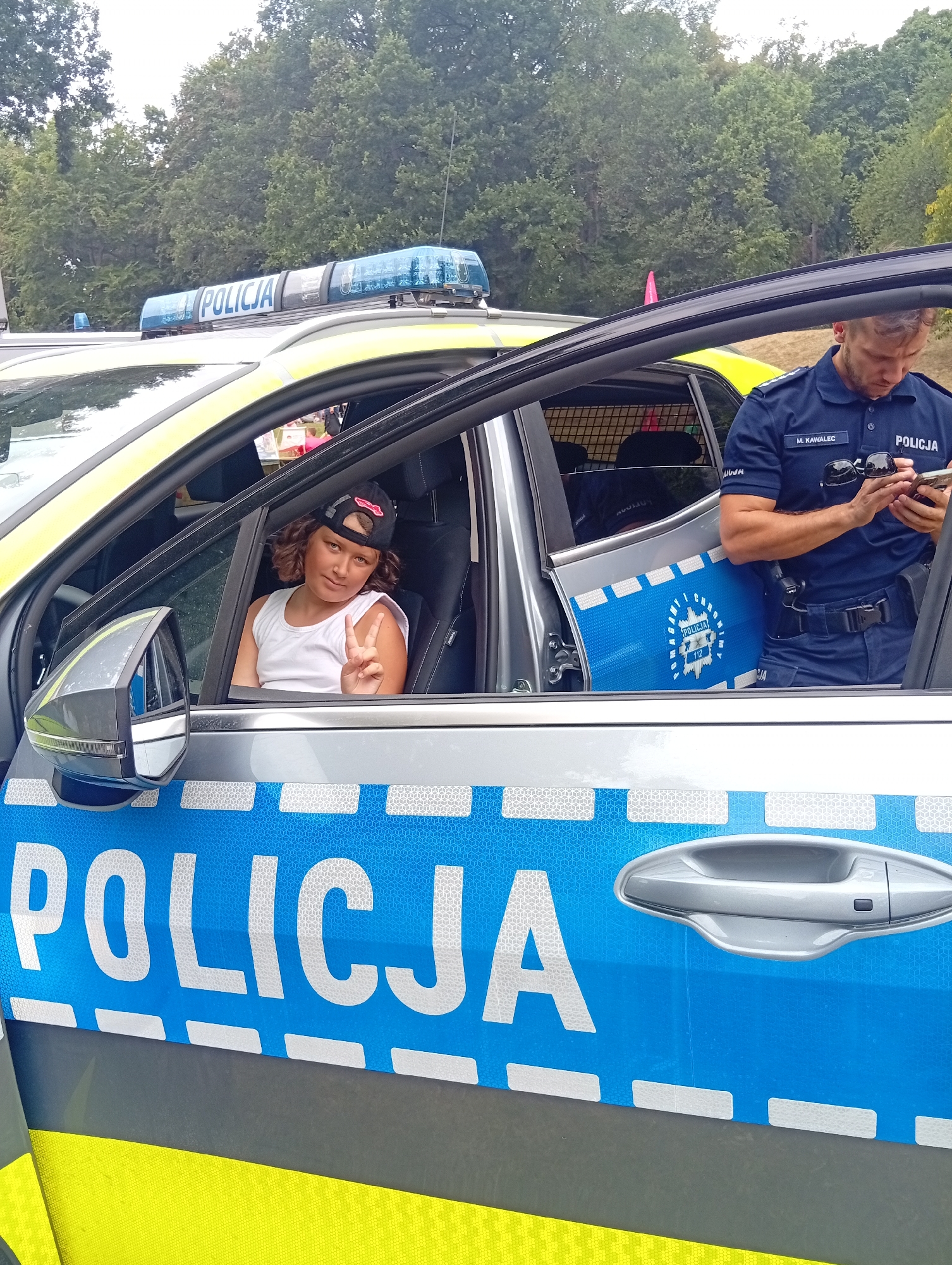 The image shows a child sitting in the driver's seat of a police car with the doors open. A police officer stands outside the car, looking at his phone. The police car has "POLICJA" written on the side. The background shows trees and a cloudy sky. The image does not provide information about the location in the city of Wrocław, Poland.