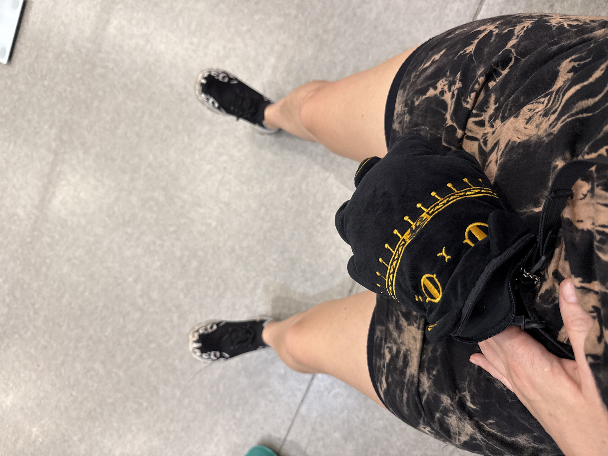 This media file is a first-person perspective shot of a person's legs and torso, viewed from above. The subject is wearing black and tan tie-dye shorts and black sneakers with a white and black patterned sole. They are holding a black plush object with yellow embroidery resembling a crown. The background is a light grey tiled floor, suggesting an indoor setting. The lighting appears to be artificial and even, with no strong shadows. The image is well-focused, with the person's legs and the plush object being the sharpest elements. There is no visible text in the image. The activity appears to be casual, possibly standing or walking in a public space such as a mall or a gym. The image is cropped tightly on the subject's lower body, with the rest of the environment not visible. The overall mood of the image is neutral and observational.