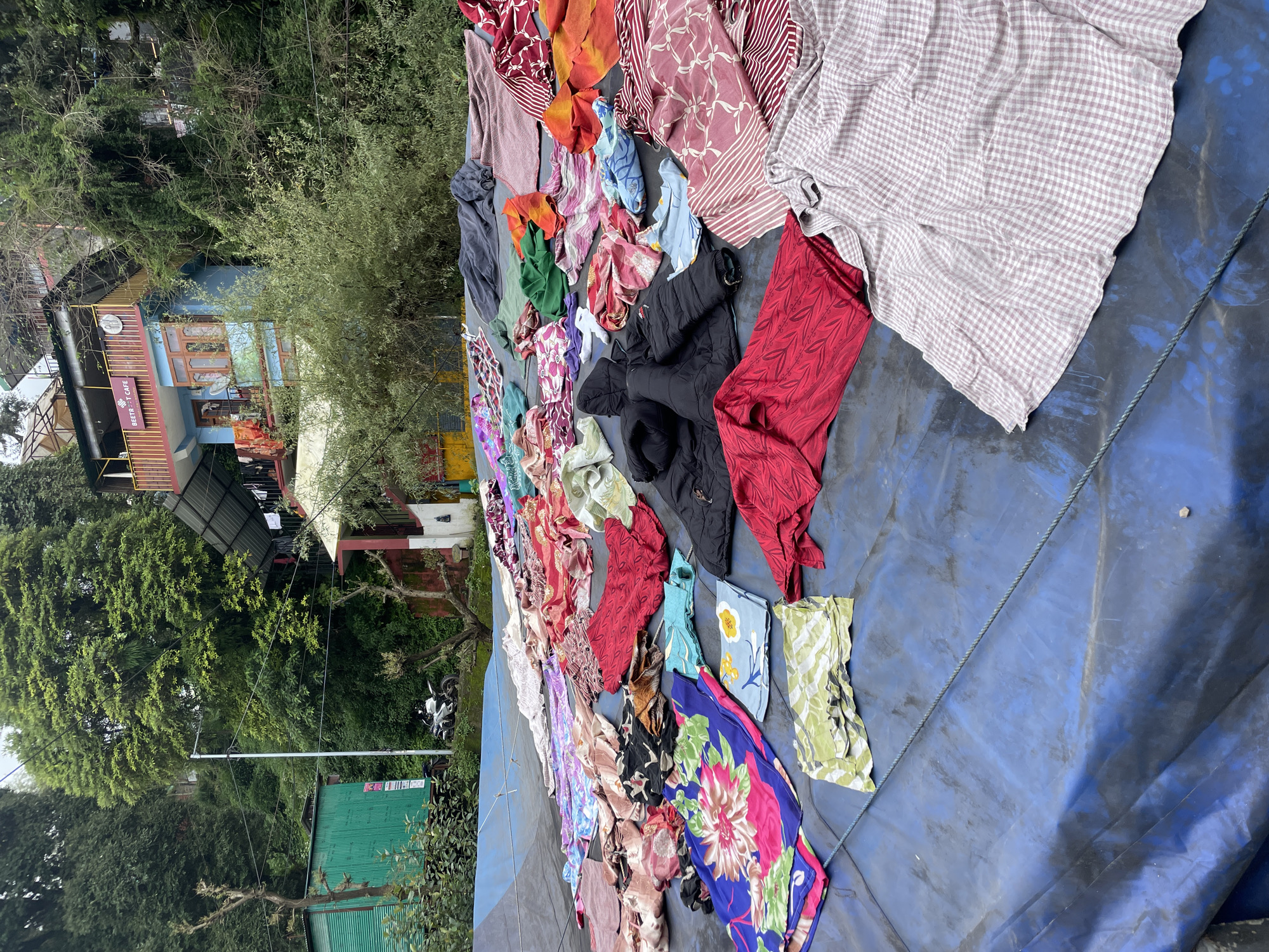 In Bhagsu, India, a blue tarp is spread on the ground, covered with an assortment of vibrantly colored clothes laid out to dry. These garments feature diverse patterns including floral prints, geometric designs, and stripes in shades of red, pink, blue, green, and orange. Amongst the clothing are two black jackets, some folded fabric squares, and a blue patterned pillowcase. In the background, a multi-story building with blue and red facades is visible, adorned with a sign that reads "BEETROOT CAFE." Lush green trees and foliage surround the building, creating a natural, hillside setting. A white motorcycle is parked near a green corrugated metal structure to the left. The scene depicts an outdoor drying area for laundry, set against the backdrop of a bustling Indian town.