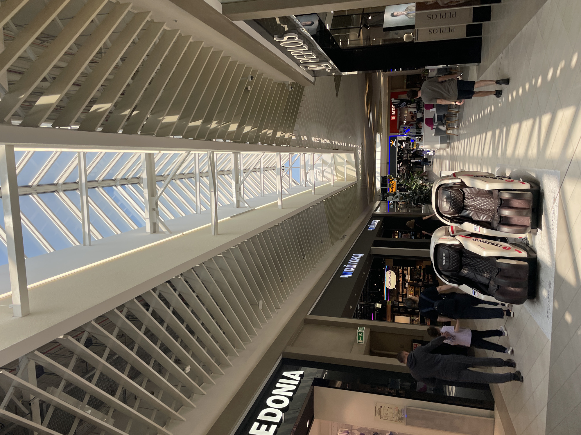 The image captures the interior of a shopping mall in Kazan, Russia. The perspective is from a slightly elevated position looking down the mall's central corridor. The ceiling is a prominent feature, composed of a grid-like structure and skylights providing natural light. The mall is lined with retail stores; recognizable brands like "MACEDONIA," "ЧИТАЙ ГОРОД," and "PEPLOS" are visible. Several people are present, some walking and others examining display items. Near the foreground, there are two massage chairs. Individuals of varying ages are present. The environment is well-lit, and the overall scene suggests a bustling commercial space.