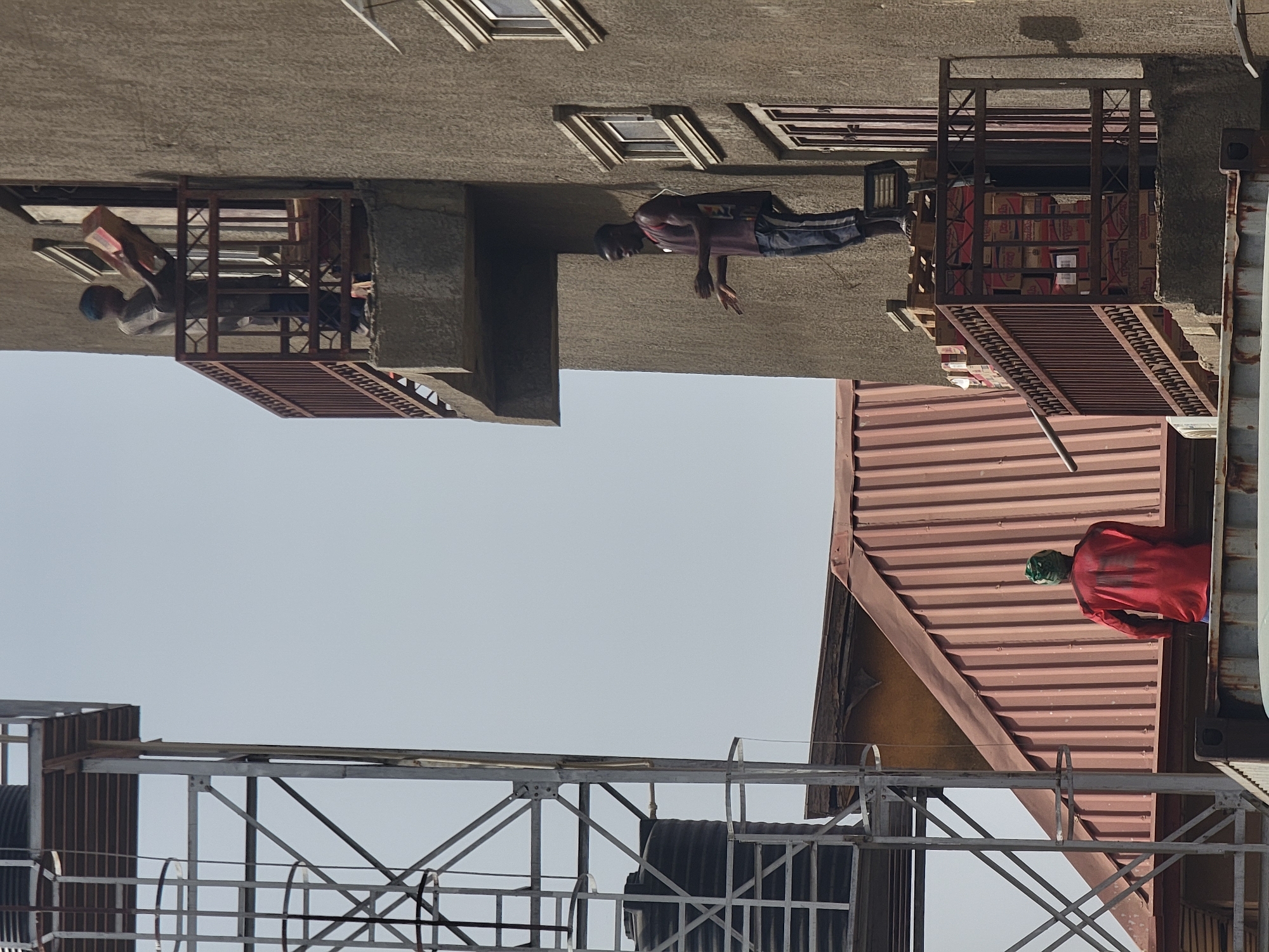 The scene shows a street scene in Maiburiji, Nigeria, with multiple buildings and people. On the upper left, a person wearing a blue head covering leans out from a balcony holding a pink object. Adjacent to this, a man in a t-shirt and shorts leans out from a higher level balcony, extending his arms. Further right, a balcony is stacked with boxes. On the right of the image, a person wearing a red shirt and green hat stands on a corrugated roof. In the foreground, a metal scaffolding structure is visible. The sky is pale and clear.