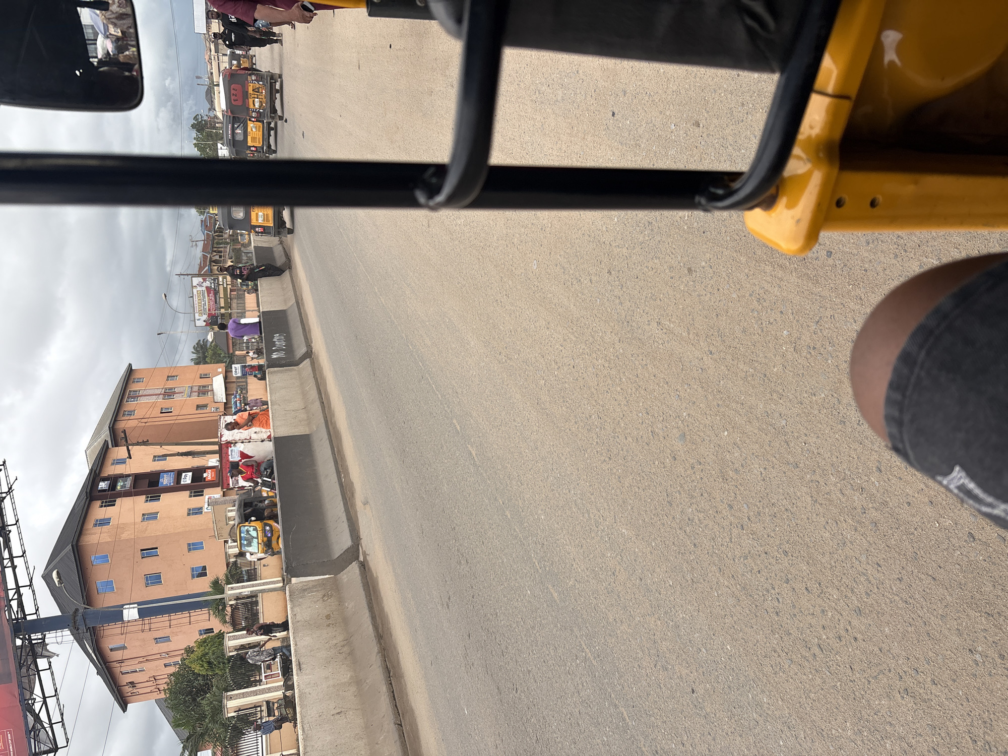 The image depicts a street scene in Onitsha, Nigeria. In the foreground, a person's legs are visible, sitting in what appears to be a yellow auto-rickshaw, commonly known as a "Keke." The Keke is positioned on a paved road. Ahead, on the right side of the road, several other auto-rickshaws are present, some in motion.
To the left of the road, a concrete barrier divides it from a sidewalk area. Along the sidewalk, a multi-story building with peach-colored walls and numerous blue-windowed floors dominates the background. Several people are visible on the sidewalk, some walking and others standing. One person wears an orange garment, and another is dressed in purple. Further down the road, more buildings and vehicles can be seen. The sky above is overcast, with visible clouds. The overall scene suggests a busy urban environment with typical street traffic and commercial activity.