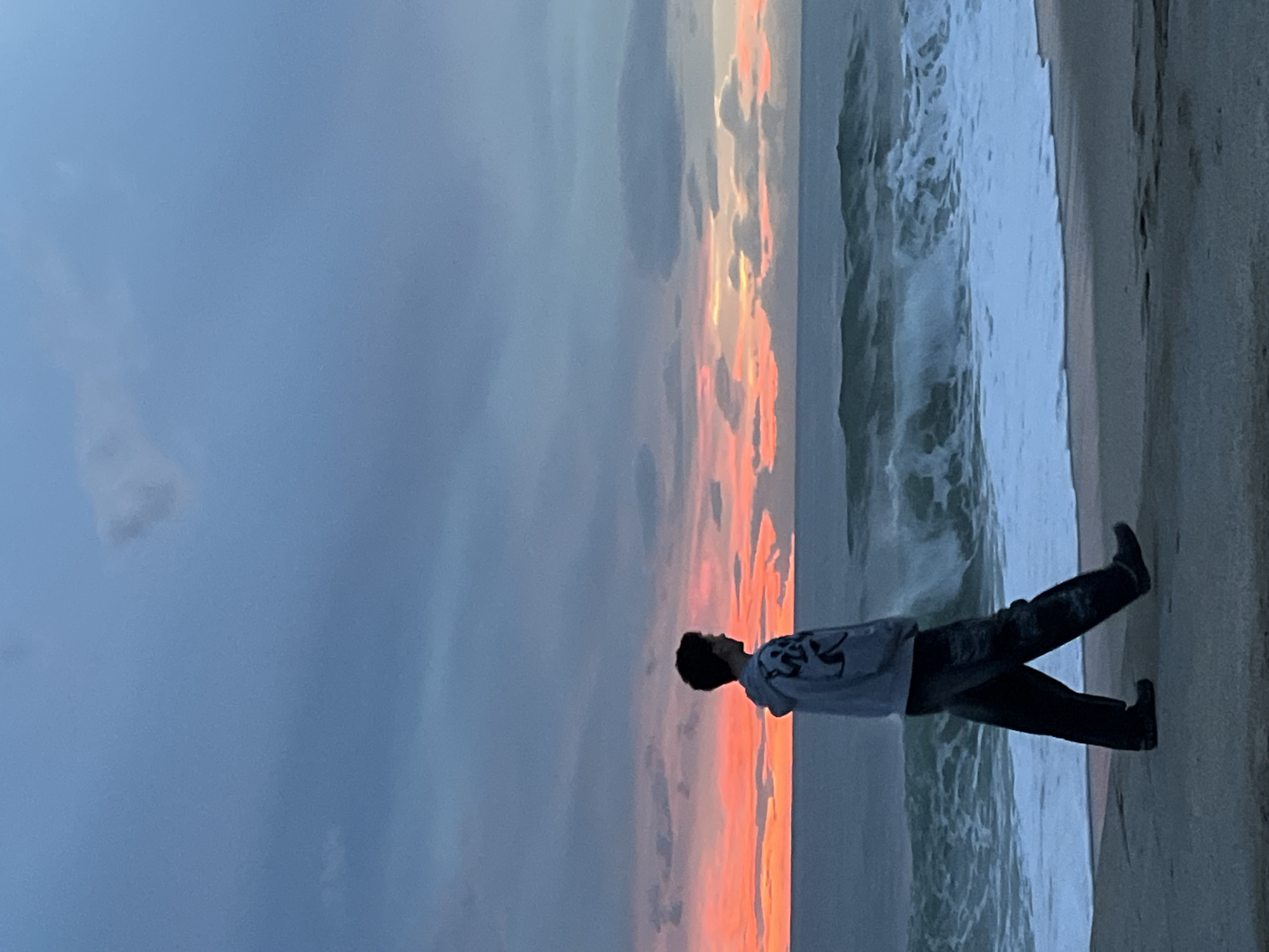 A person stands on a beach in Alanya, Turkey, facing the ocean. The sky is a gradient of grey-blue to orange-pink, suggesting sunrise or sunset. Waves are crashing on the shore. The person is wearing dark pants and a light-colored shirt.