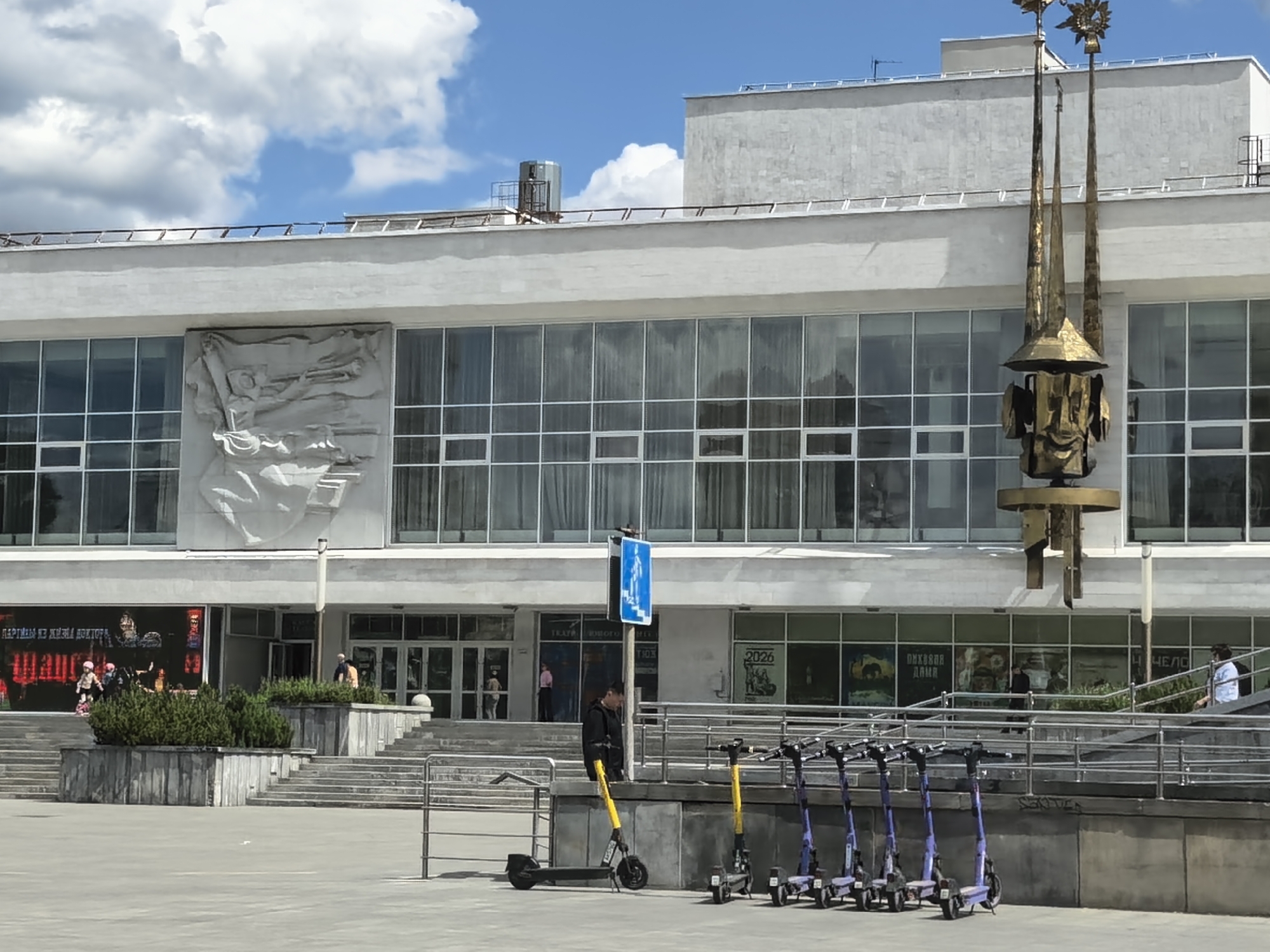 The image depicts the facade of a building in an urban environment, likely a public space, in Unknown, Unknown. The building has a large, rectangular design with numerous windows and a bas-relief sculpture on the upper part of its exterior. In front of the building, there is a row of parked electric scooters, some yellow and some purple, with a man standing near one of them. Pedestrians are visible entering and exiting the building. The sky is partly cloudy. A sculpture is next to the right-hand side of the building.