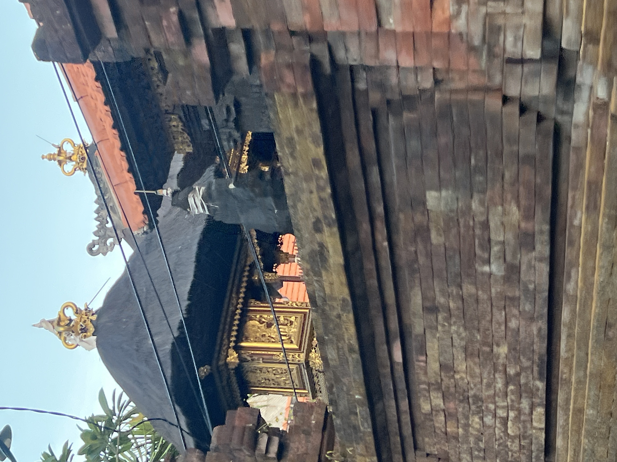 This is a medium-shot, vertical, outdoor, eye-level photograph of a traditional Balinese temple structure in Kuta, Indonesia. The foreground is dominated by a rough, weathered brick wall constructed from reddish-brown bricks. Above this wall, a tiered temple building is visible, featuring a dark, thatched roof with intricate golden decorations. The roof has a distinctive conical shape, topped with a golden, crown-like ornament. Adjacent to this, another section of the temple has an orange-tiled roof. The temple's facade is adorned with detailed golden carvings and what appear to be religious figures. A portion of lush green foliage can be seen to the left of the temple, suggesting a tropical environment. The sky above is a pale, clear blue. The image is slightly blurred, indicating motion or a shallow depth of field. There are no people visible in the frame.