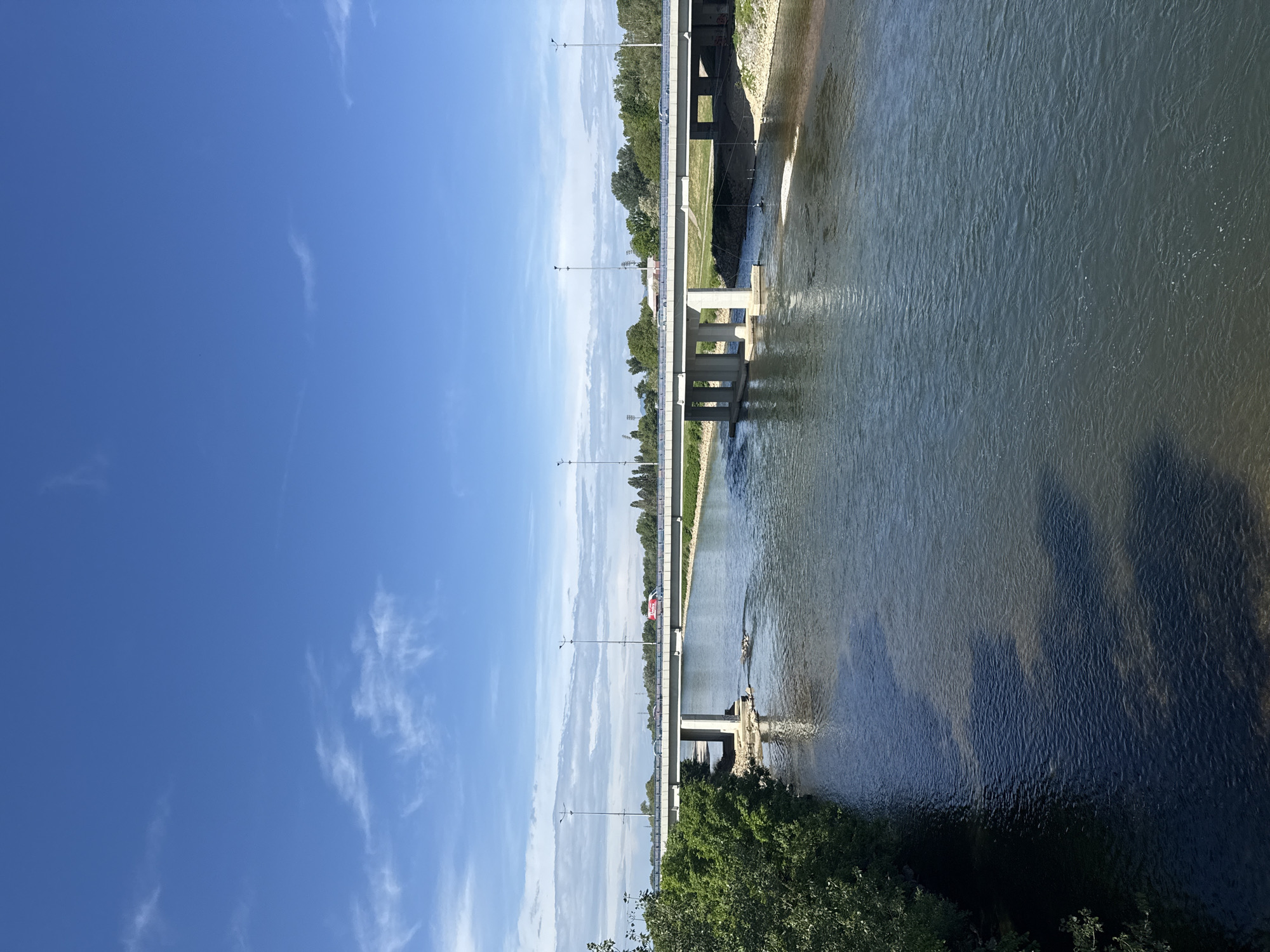 The image shows a bridge spanning a body of water. The sky is blue with scattered clouds. On the bridge, there are lampposts and vegetation. Below the bridge, the water appears to be flowing. The scene is likely in the City of Zagreb, Croatia, given the presence of a bridge over water, which is common in the city.