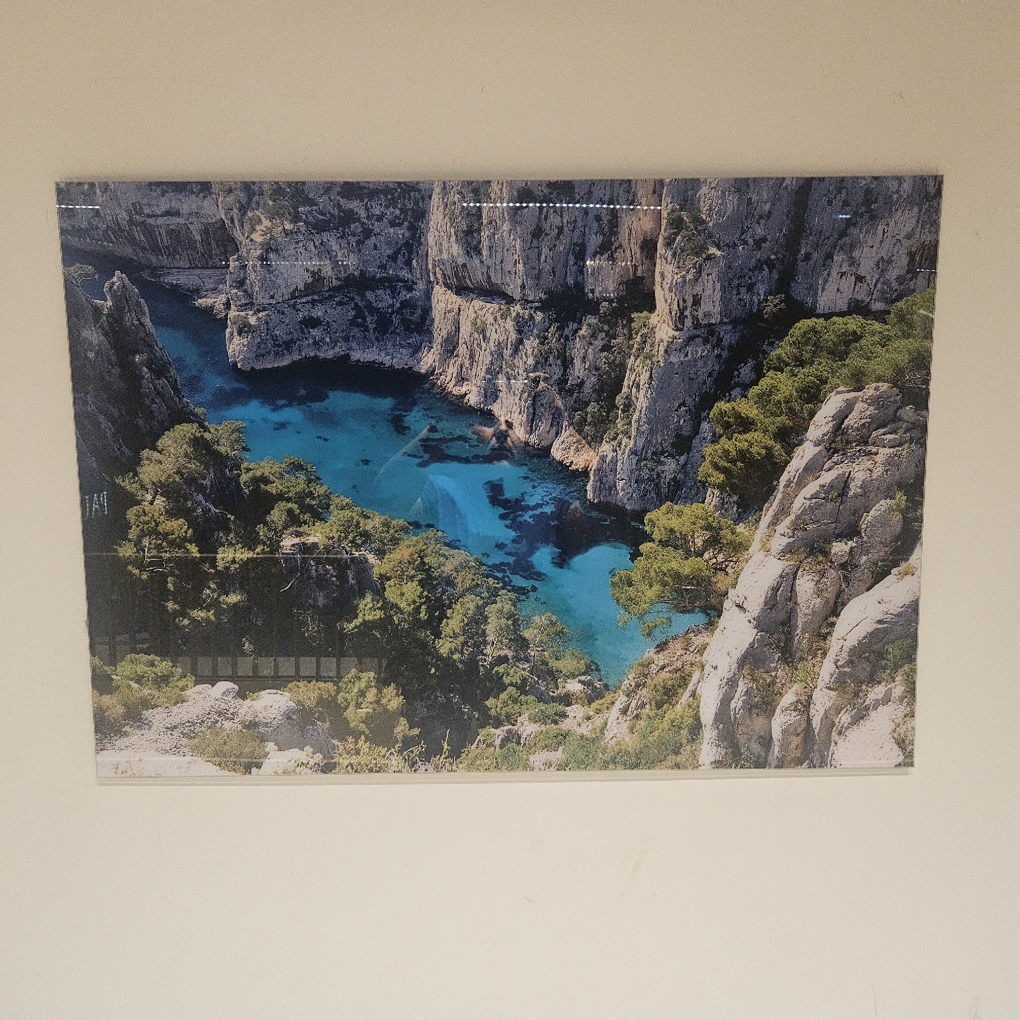 The image shows a framed print of a scenic coastal landscape. The print depicts a narrow, turquoise bay surrounded by towering, rugged, grey cliffs. Lush green trees and vegetation cling to the rocky slopes, adding a vibrant contrast to the stone and water. The water itself is clear and appears to be shallow in places, revealing darker submerged rocks and seabed. The overall impression is one of a secluded and naturally beautiful cove, likely a natural phenomenon in a coastal region. The print is mounted on a plain, light-colored wall. The location in the city of Lançon-Provence, France is not visible in the image itself, and therefore cannot be determined from the provided visual information. There are no people, animals, or specific objects or actions that suggest a narrative beyond the depiction of the landscape.